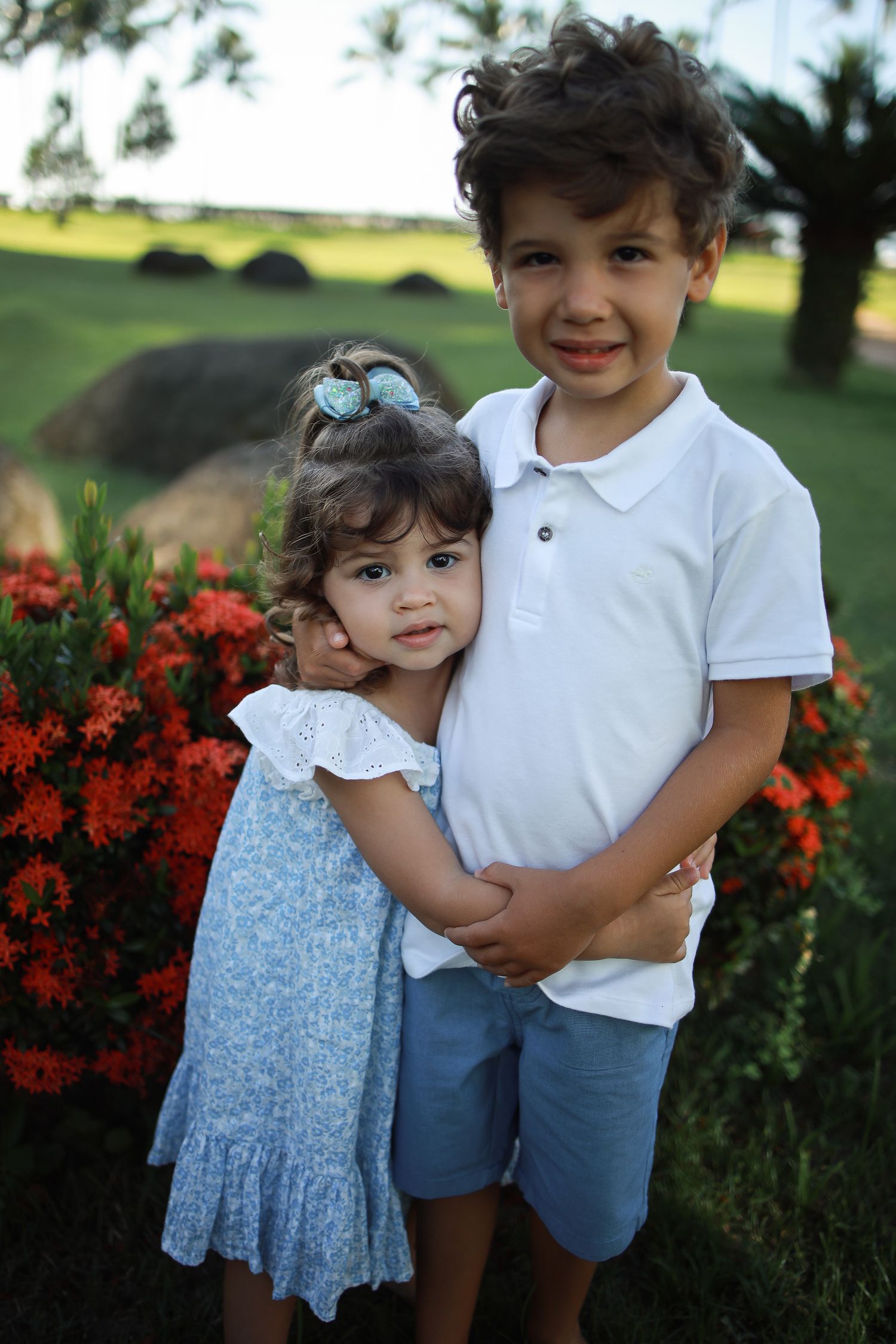Two children posing together near blooming red flowers in garden setting.