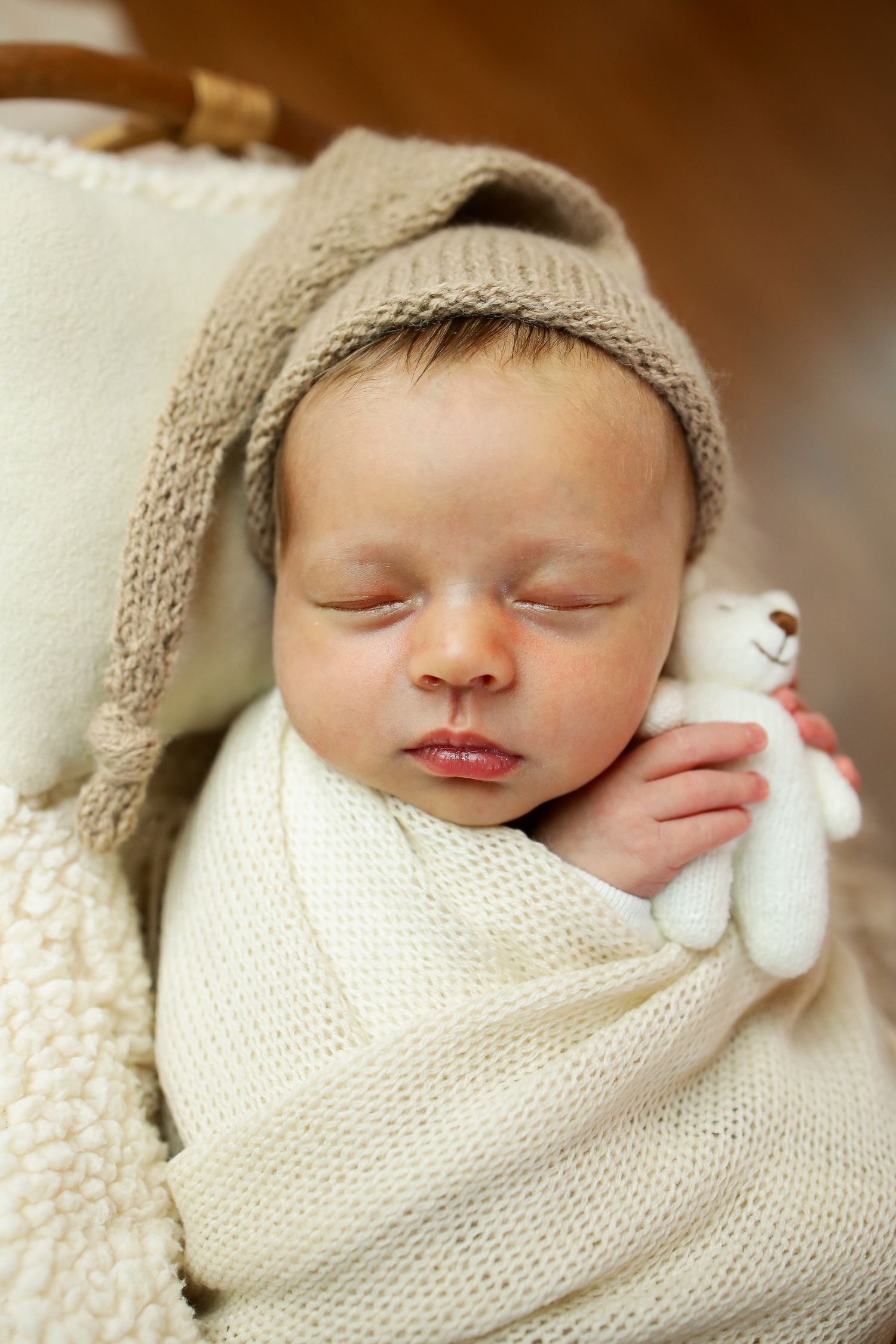 Peaceful sleeping newborn wrapped in cozy cream knit blanket and bonnet holding small stuffed toy.