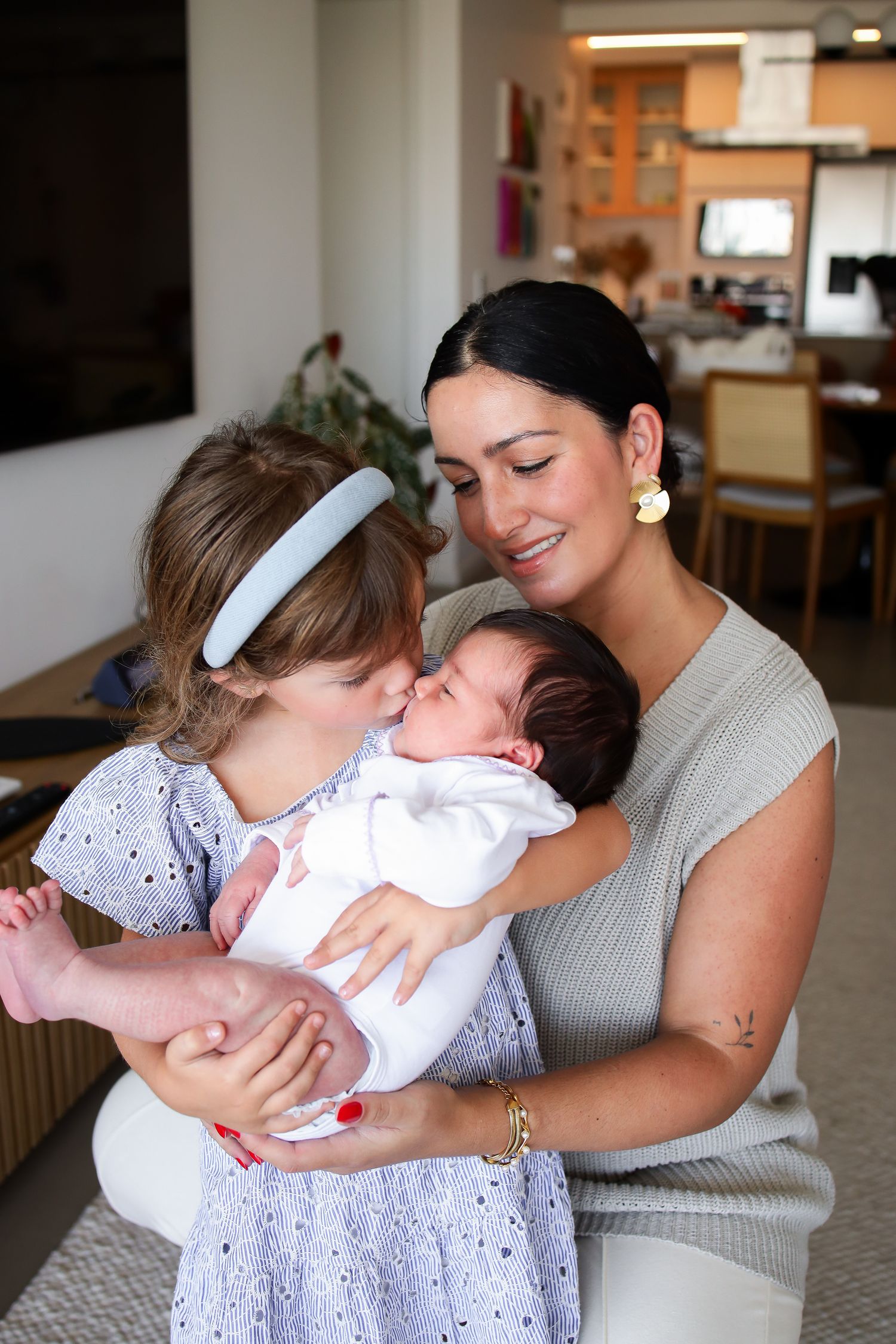 A tender family moment shows a young girl in white dress loving gazing at a newborn baby being held in a home setting.