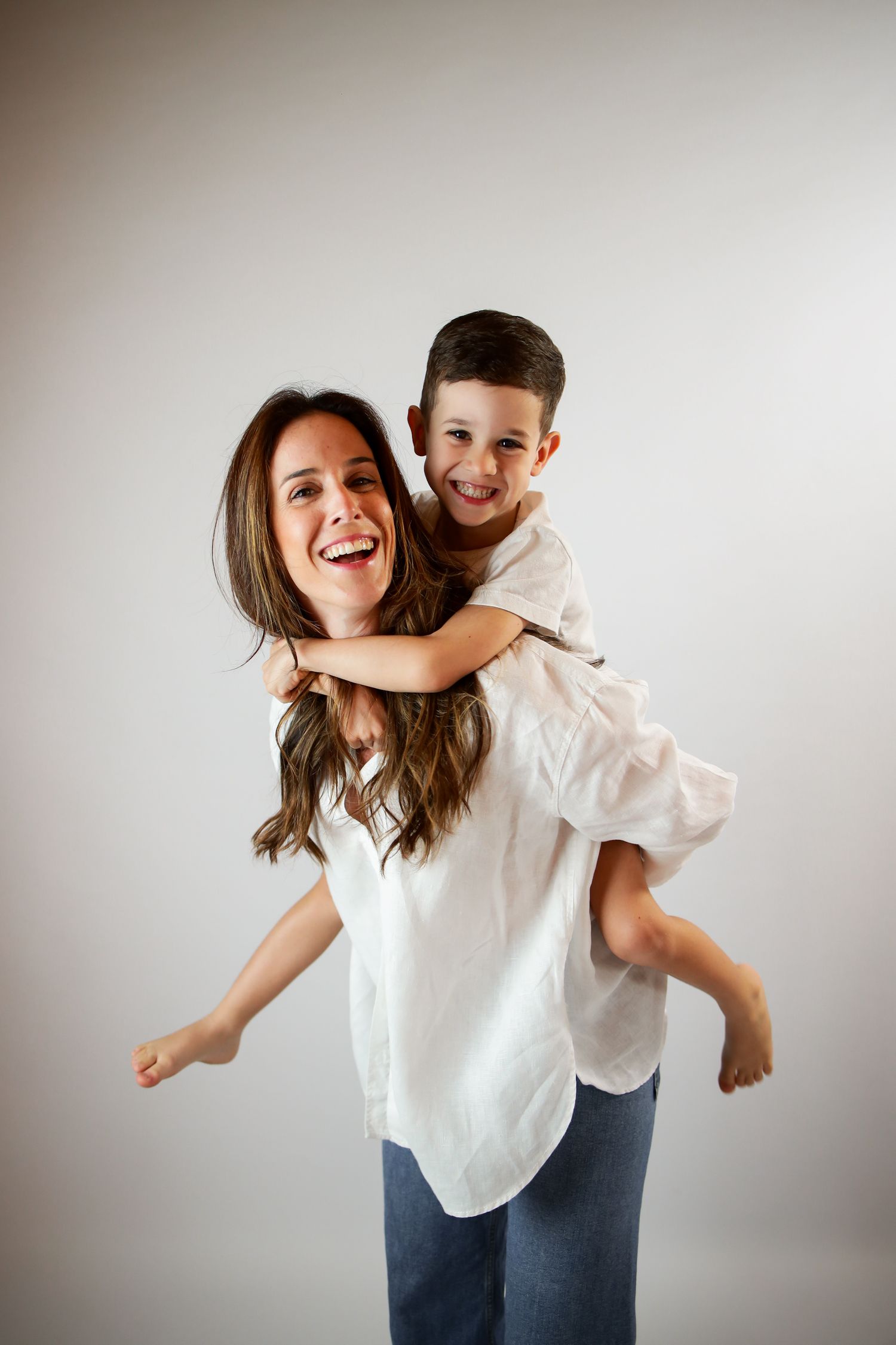 A joyful mother and son share a playful piggyback ride moment against a neutral background.