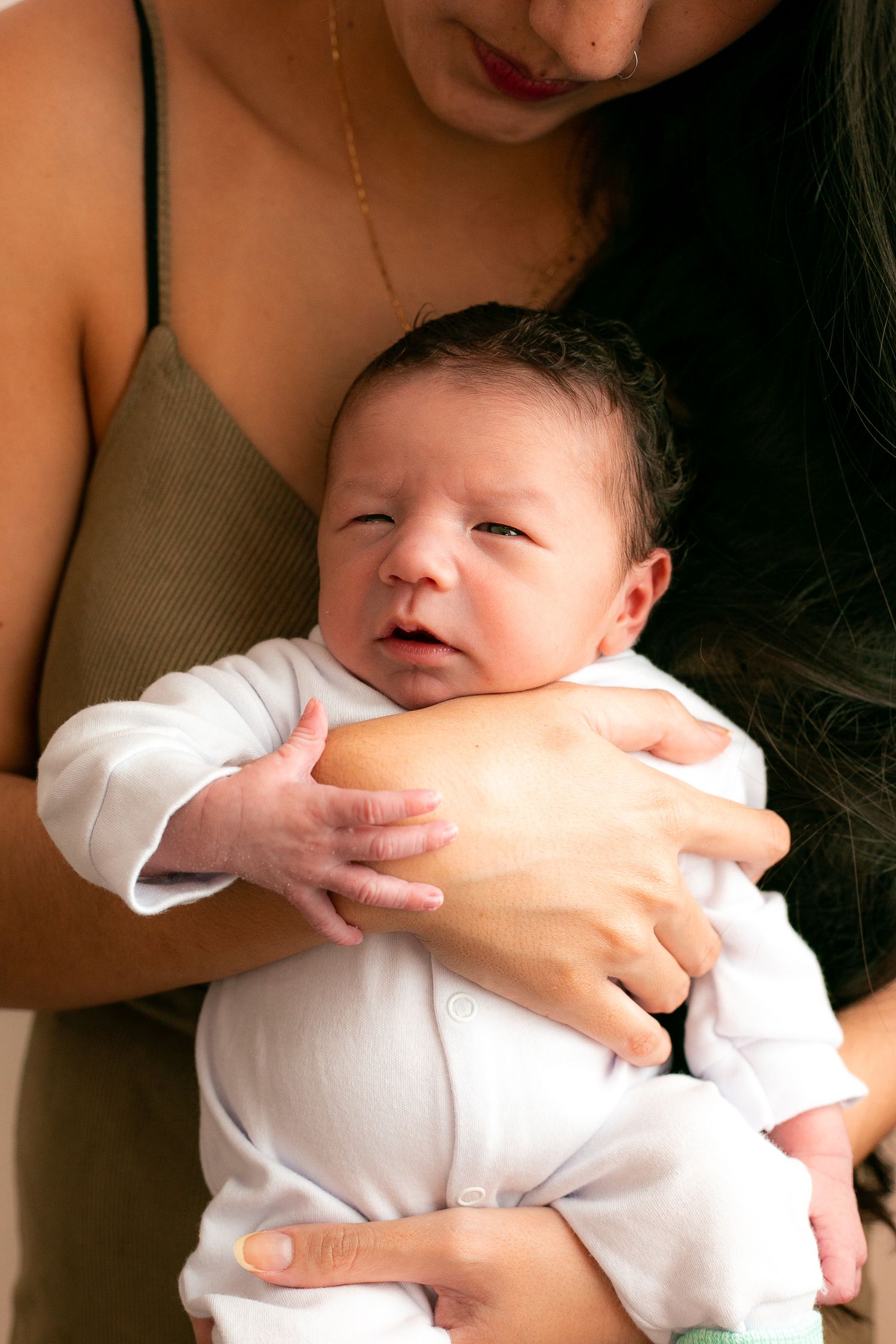 A newborn baby wearing a white onesie is held in loving arms while making an expressive face.