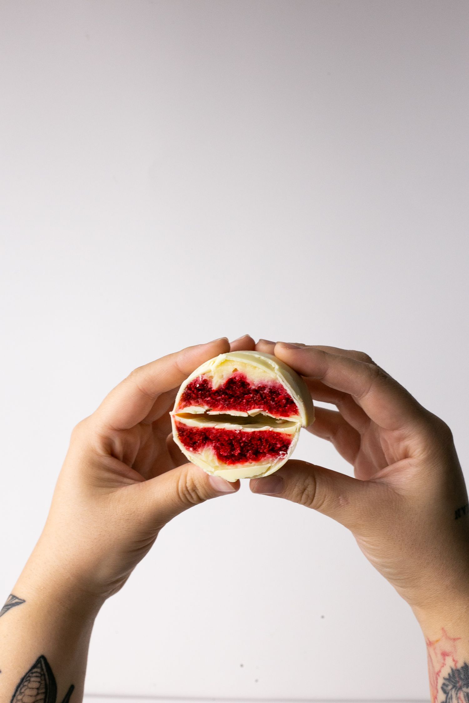 Hands holding a split cookie filled with bright red jam against a light gray background.