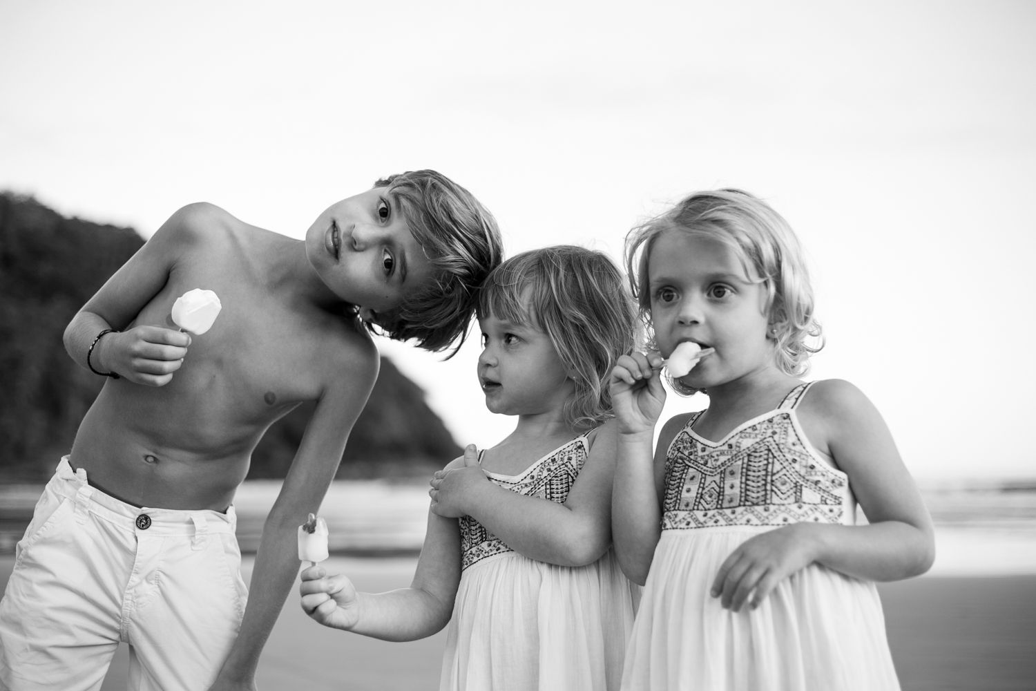 Three young children in casual summer outfits enjoying popsicles together at the beach during sunset.