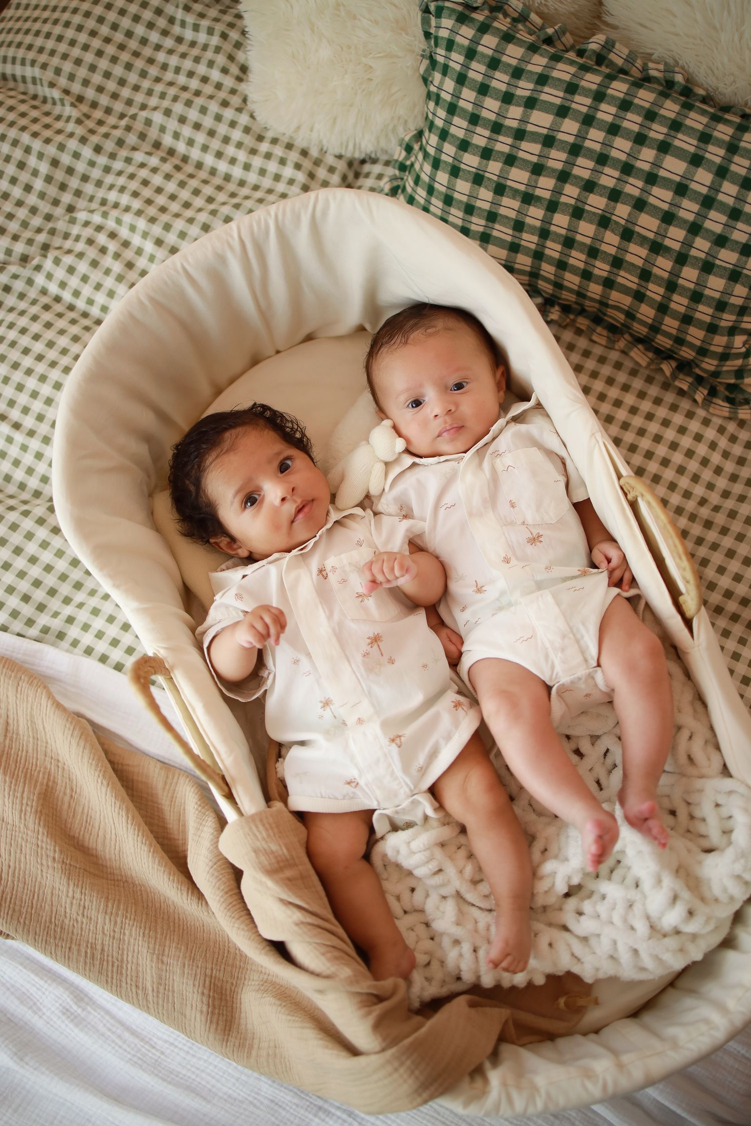 Two infants wearing white outfits lie together in a cozy cream-colored bassinet with gingham pillows in the background.
