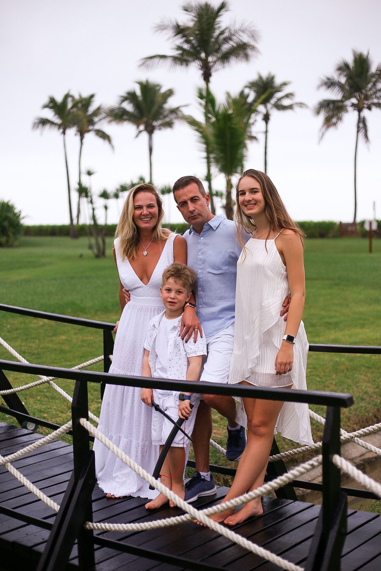 Family in white summer outfits poses together on wooden deck with tropical palm trees in background.
