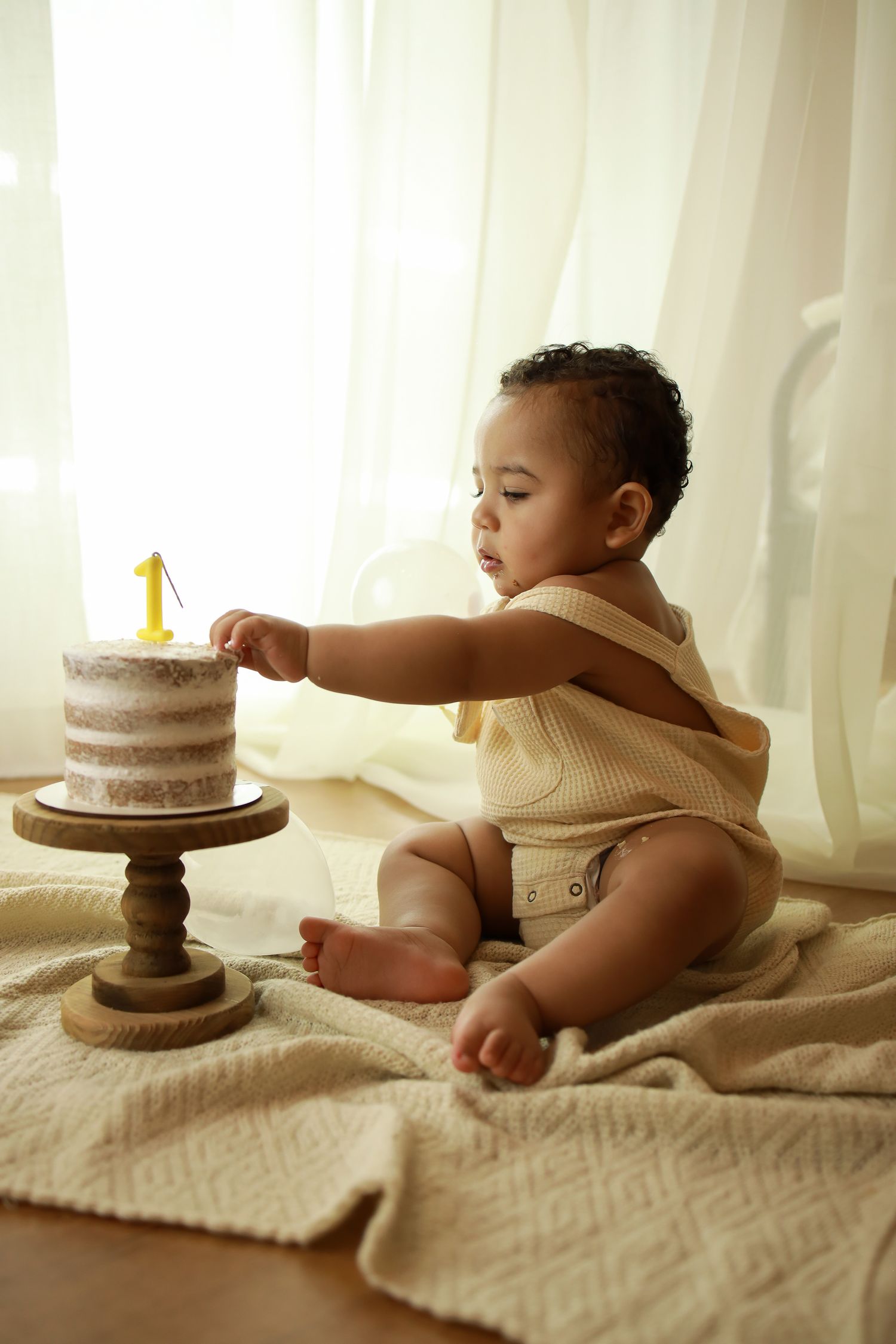 Baby in cream-colored outfit reaching for small birthday cake on wooden stand.
