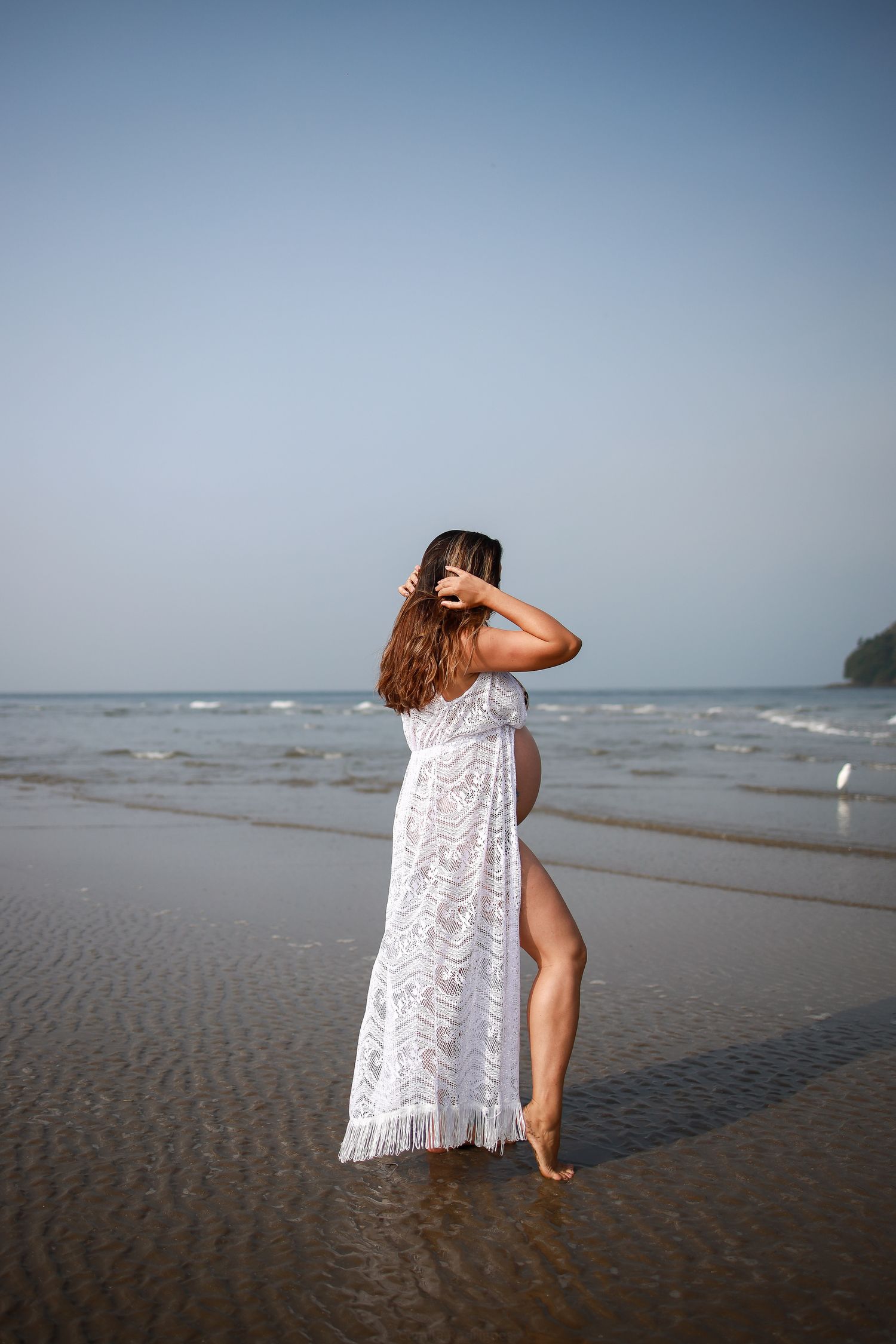 A figure in a flowing white lace dress poses on a misty beach shoreline at sunset.