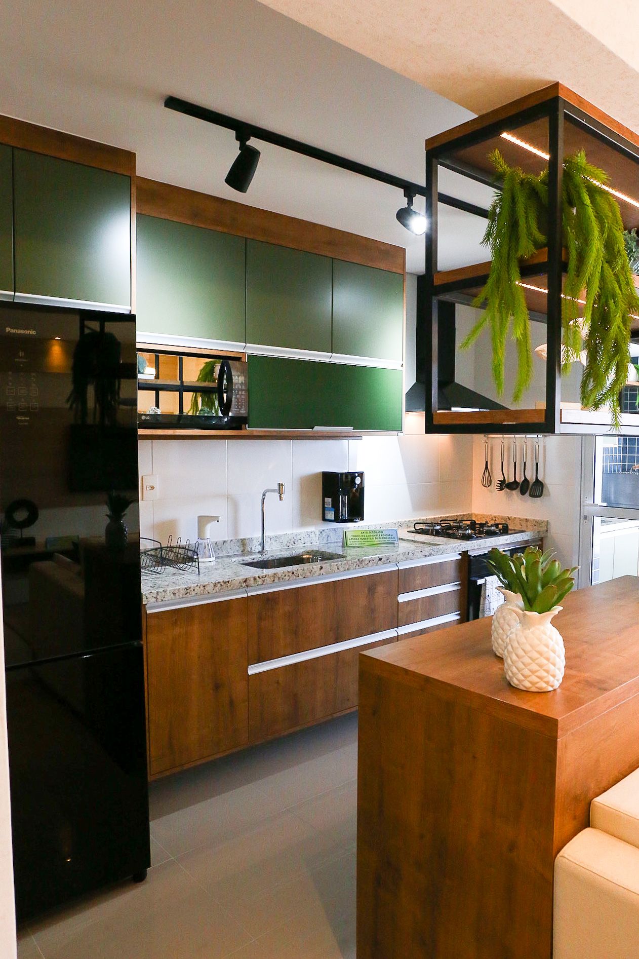 Modern kitchen design with green upper cabinets, wooden lower cabinets, black appliances and hanging plants in a compact space.