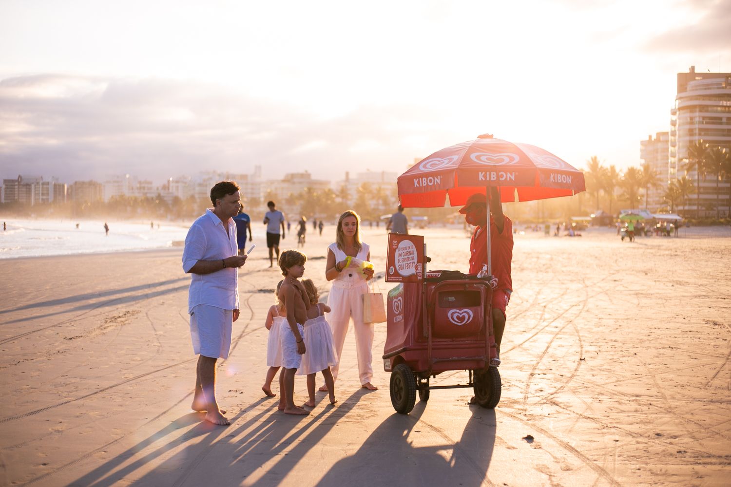 A group walks along a beach at sunset with a red umbrella cart on a warm summer evening.