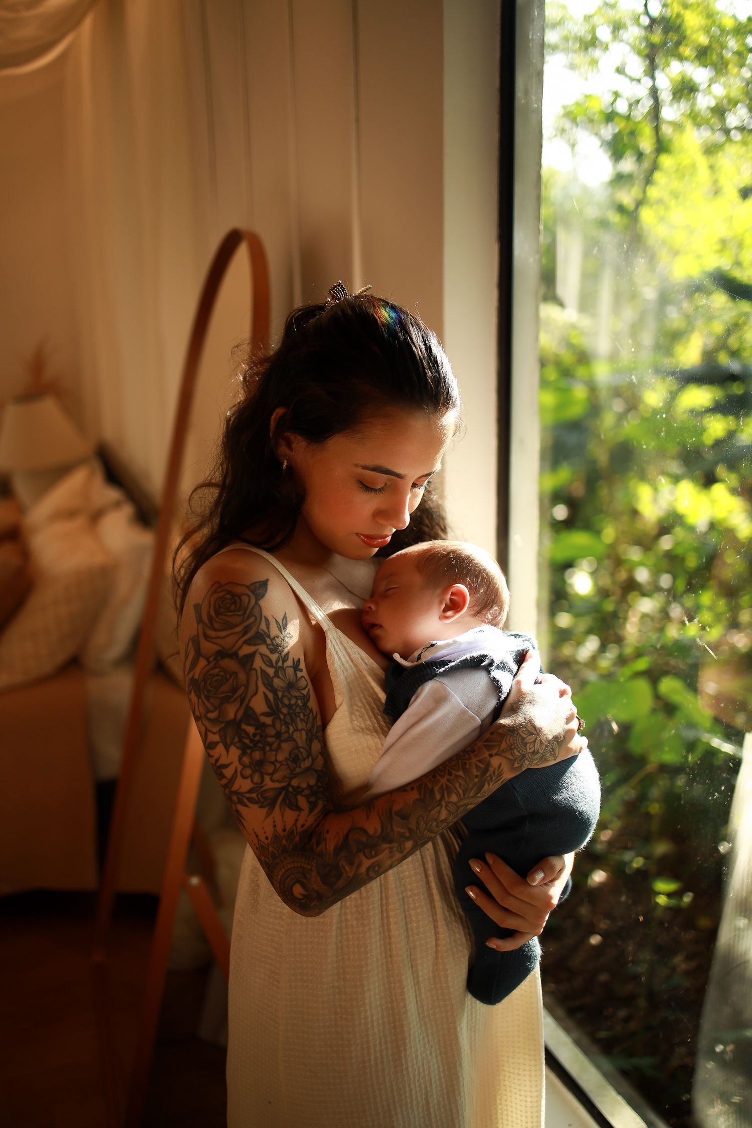 A tender moment by a sunlit window shows a parent in a white dress with tattooed arms cradling a sleeping baby.