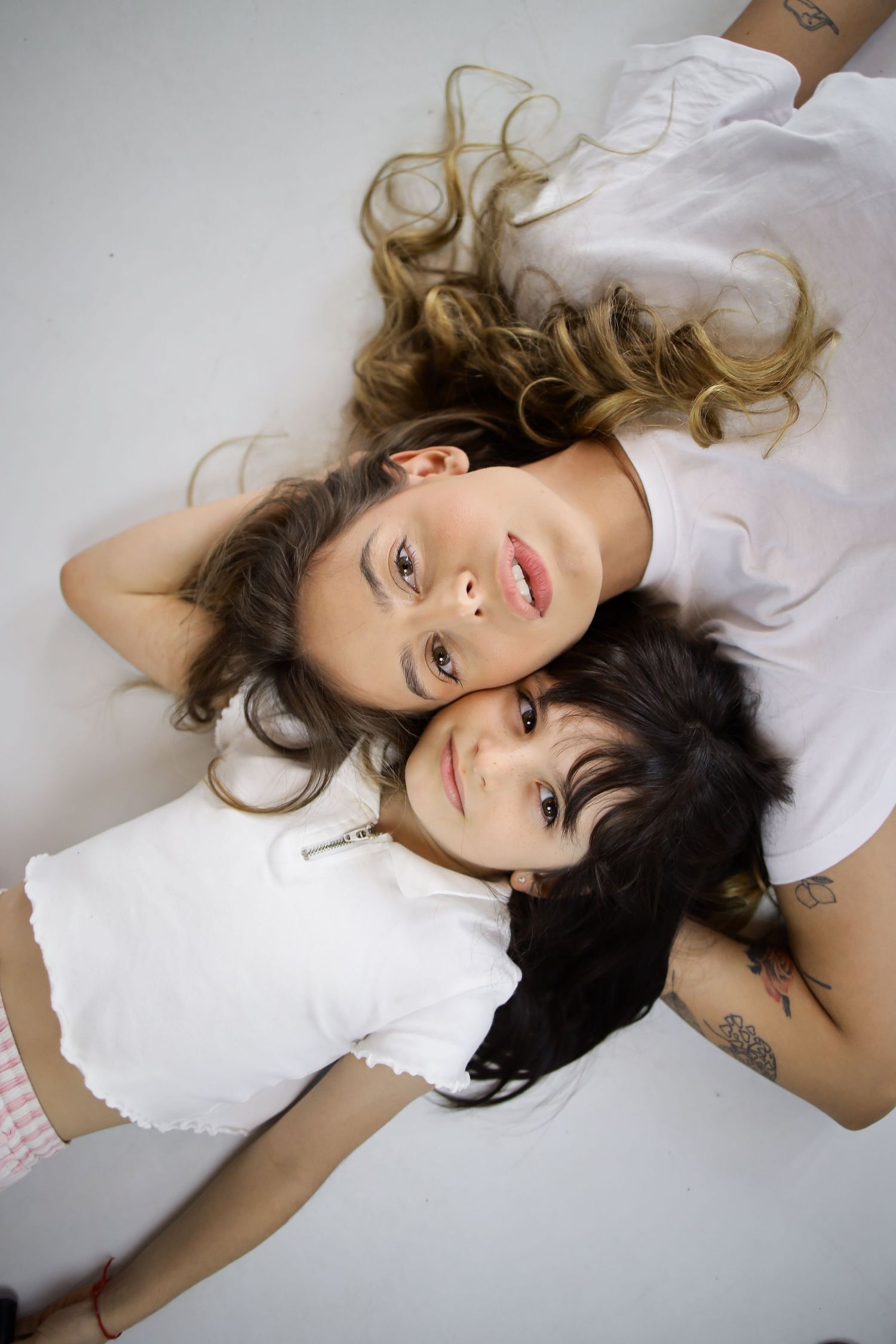 Two young women lying down together, wearing white tops, with long hair spread out, looking up at the camera.