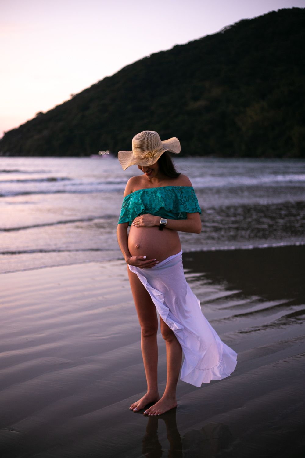Expectant mother in teal off-shoulder top and white skirt stands on beach at sunset, cradling baby bump.