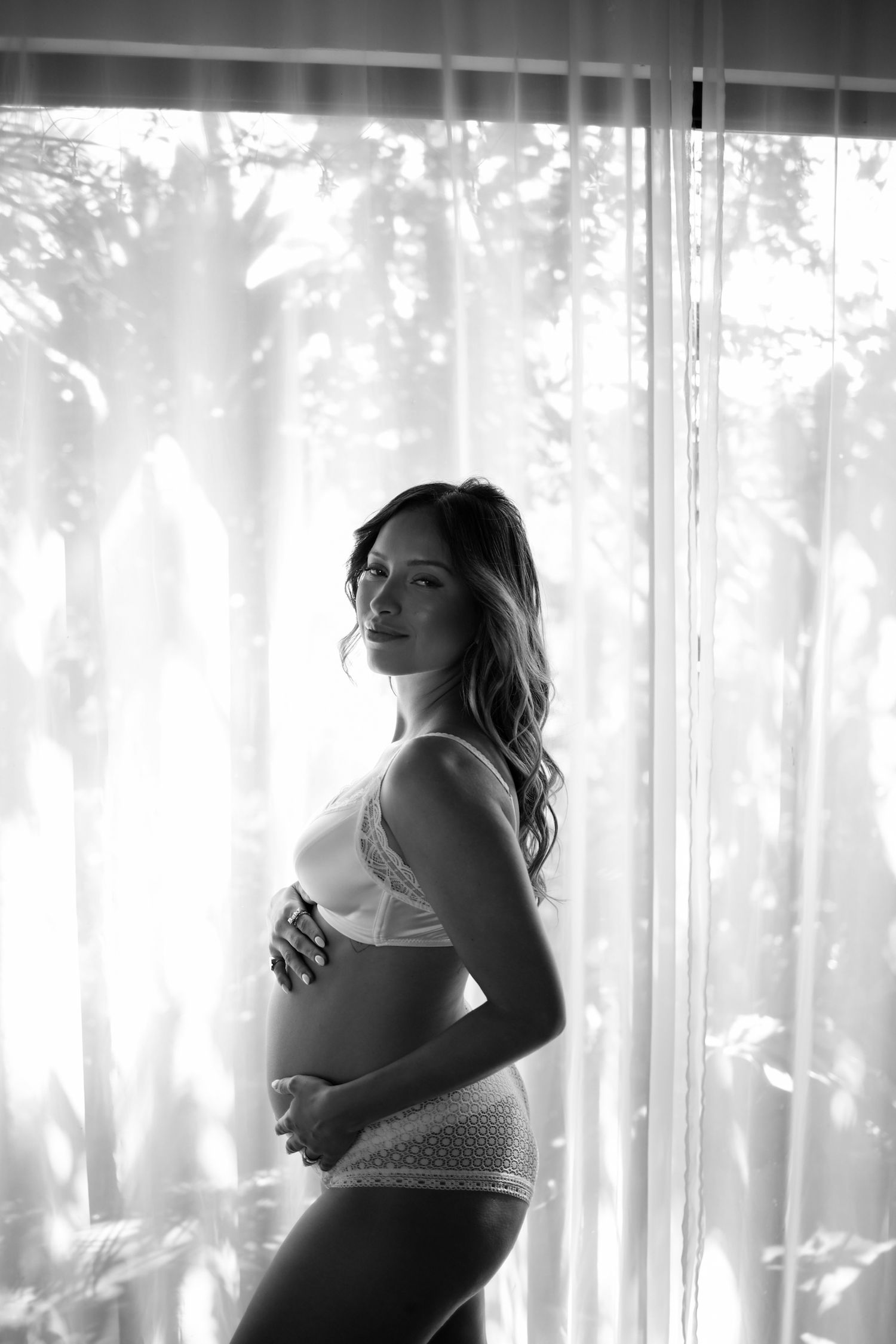 A black and white silhouette of an expectant mother by a window with sunlight filtering through sheer curtains.