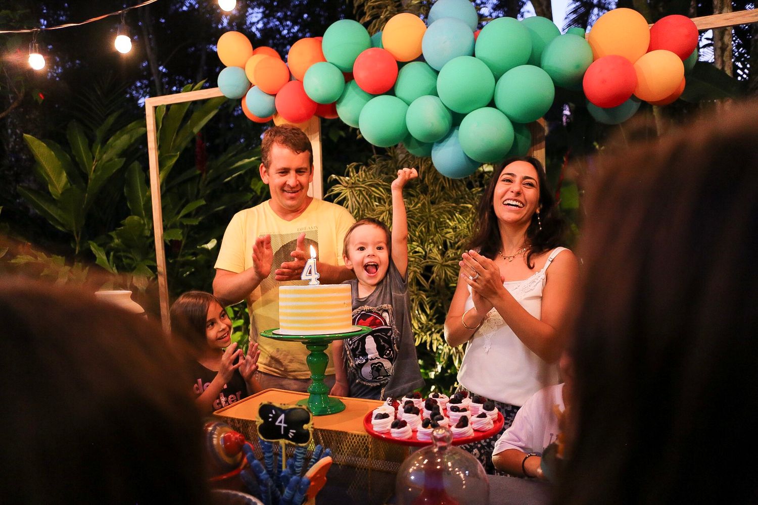 Colorful balloon arch decorates an outdoor birthday party celebration with cake and desserts on display at night.