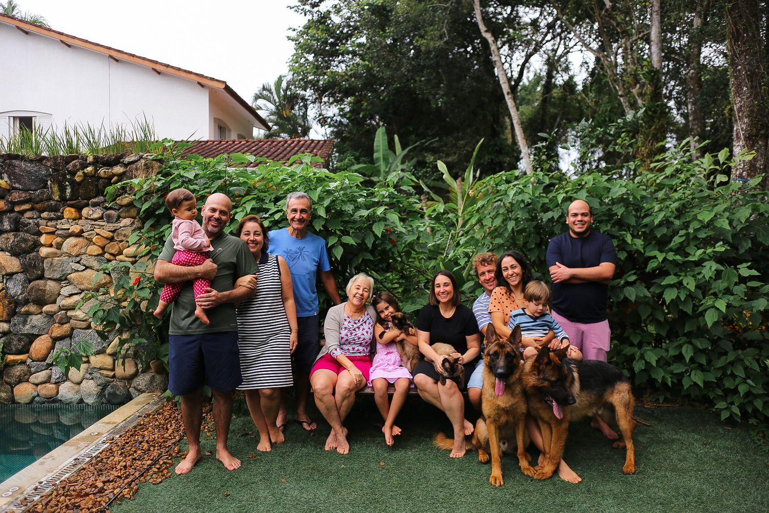 A group gathers in a backyard garden with two dogs, standing by a stone wall and lush greenery.