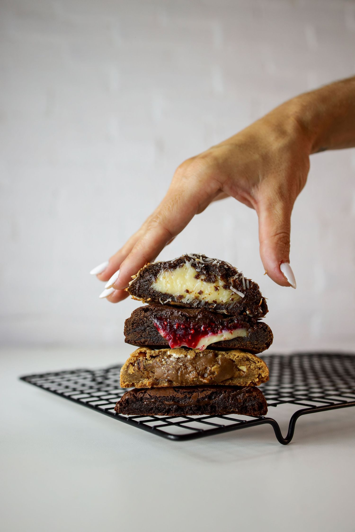 A hand reaches for a stacked chocolate dessert sandwich with cream and berry filling on a polka dot cooling rack.