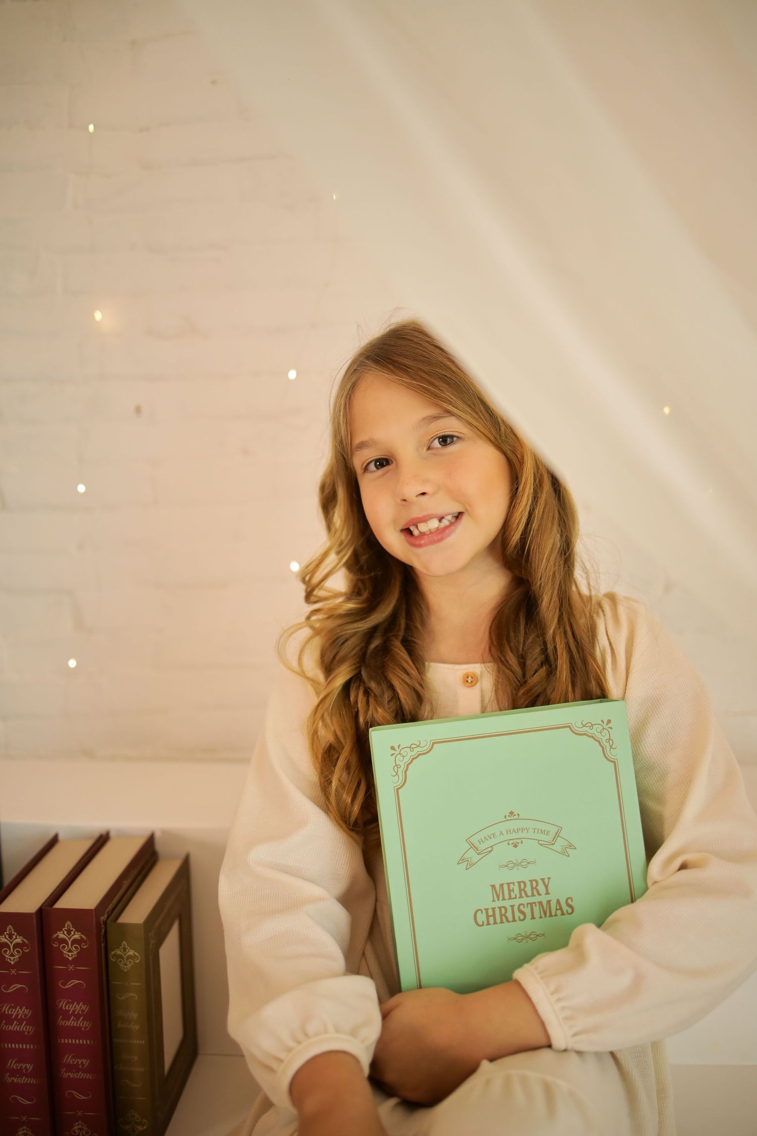 A child in a white sweater poses with a mint green book against a soft white backdrop with twinkling lights.