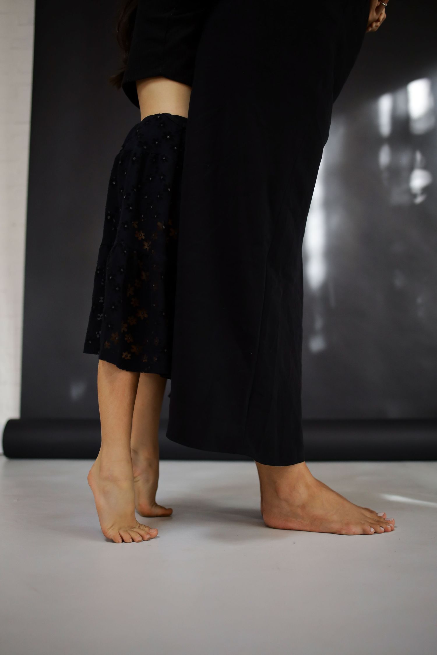 Barefoot person standing on white floor wearing black skirt and pants in a studio setting.