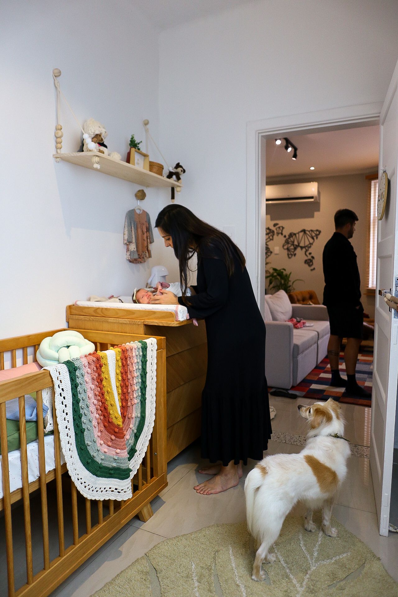 Person in black dress changing a baby at wooden changing table with crib beside them while dog watches in hallway.