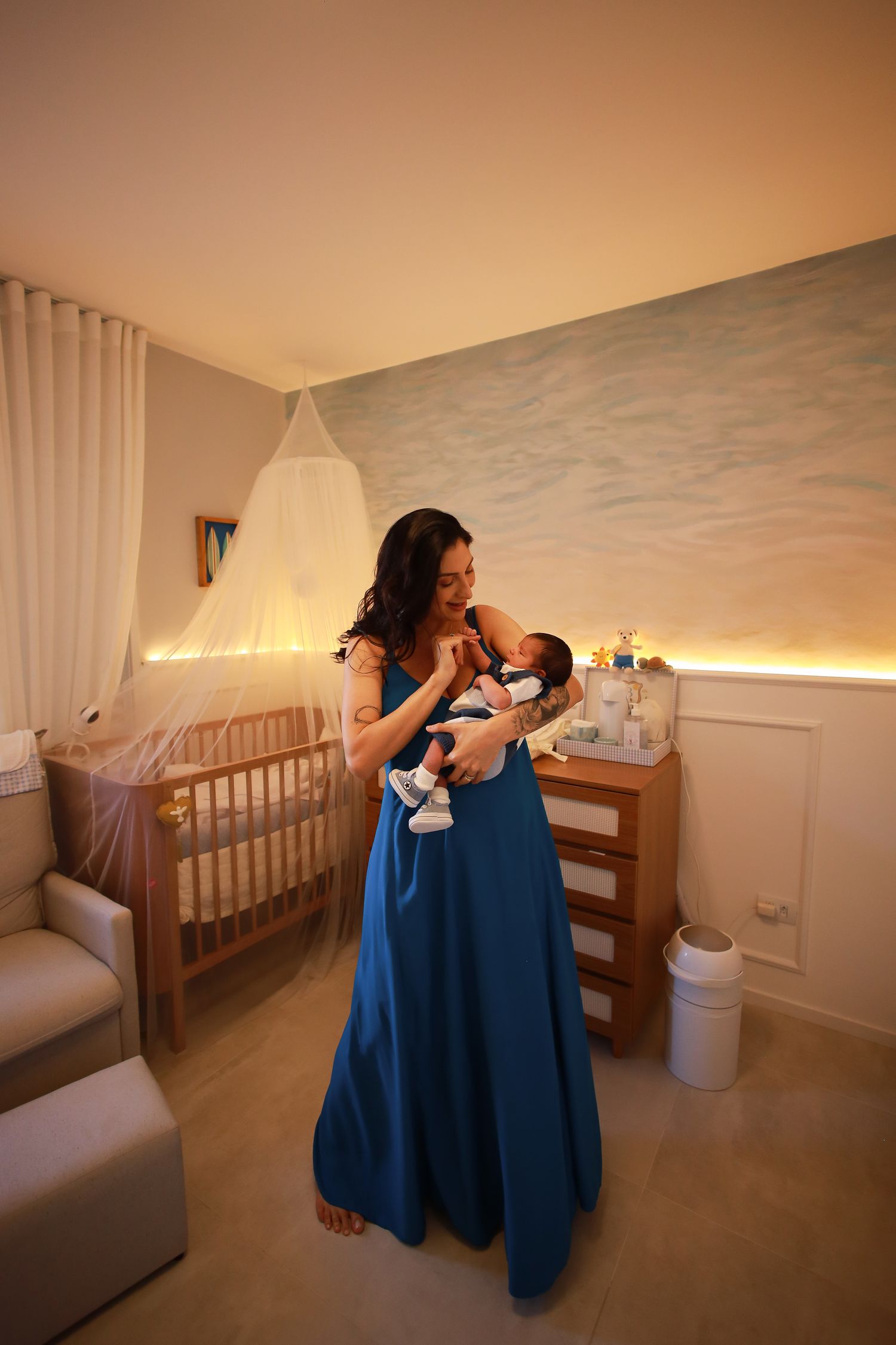 A nursery room with warm lighting features a wooden crib, curtains, and a person in a blue dress holding a baby.