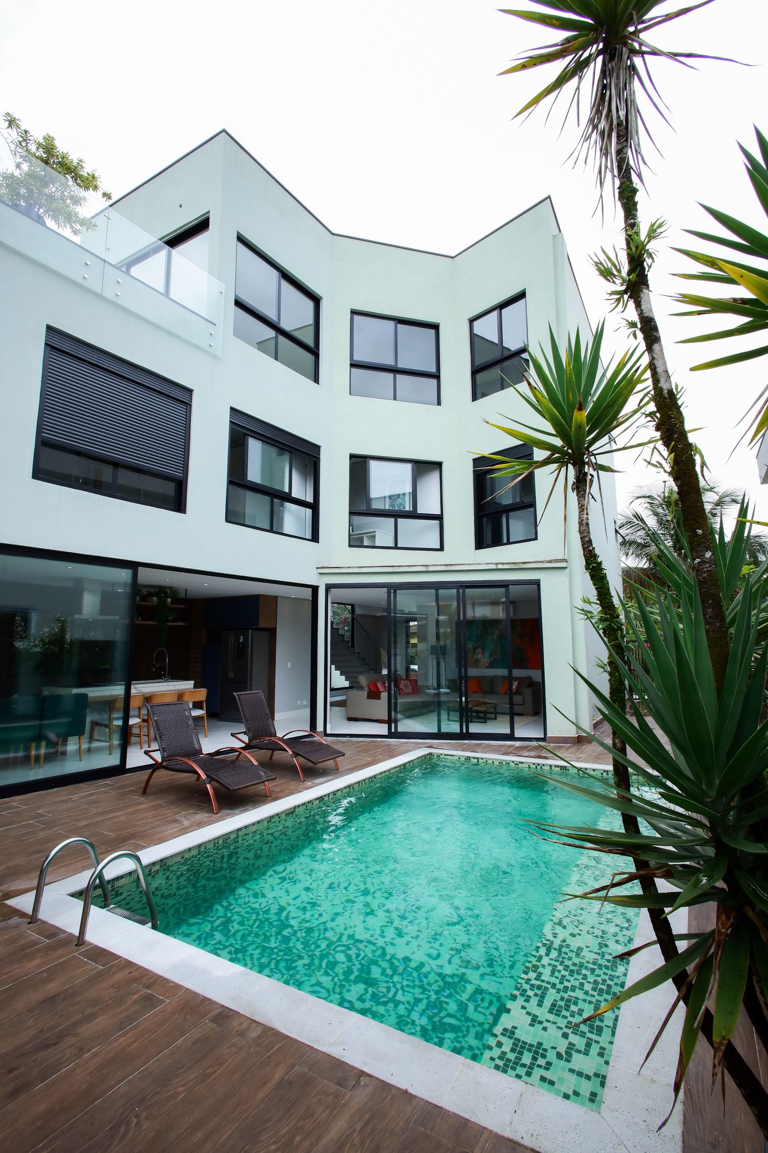 Modern white house with angular architecture features a turquoise swimming pool and wooden deck surrounded by tropical plants.
