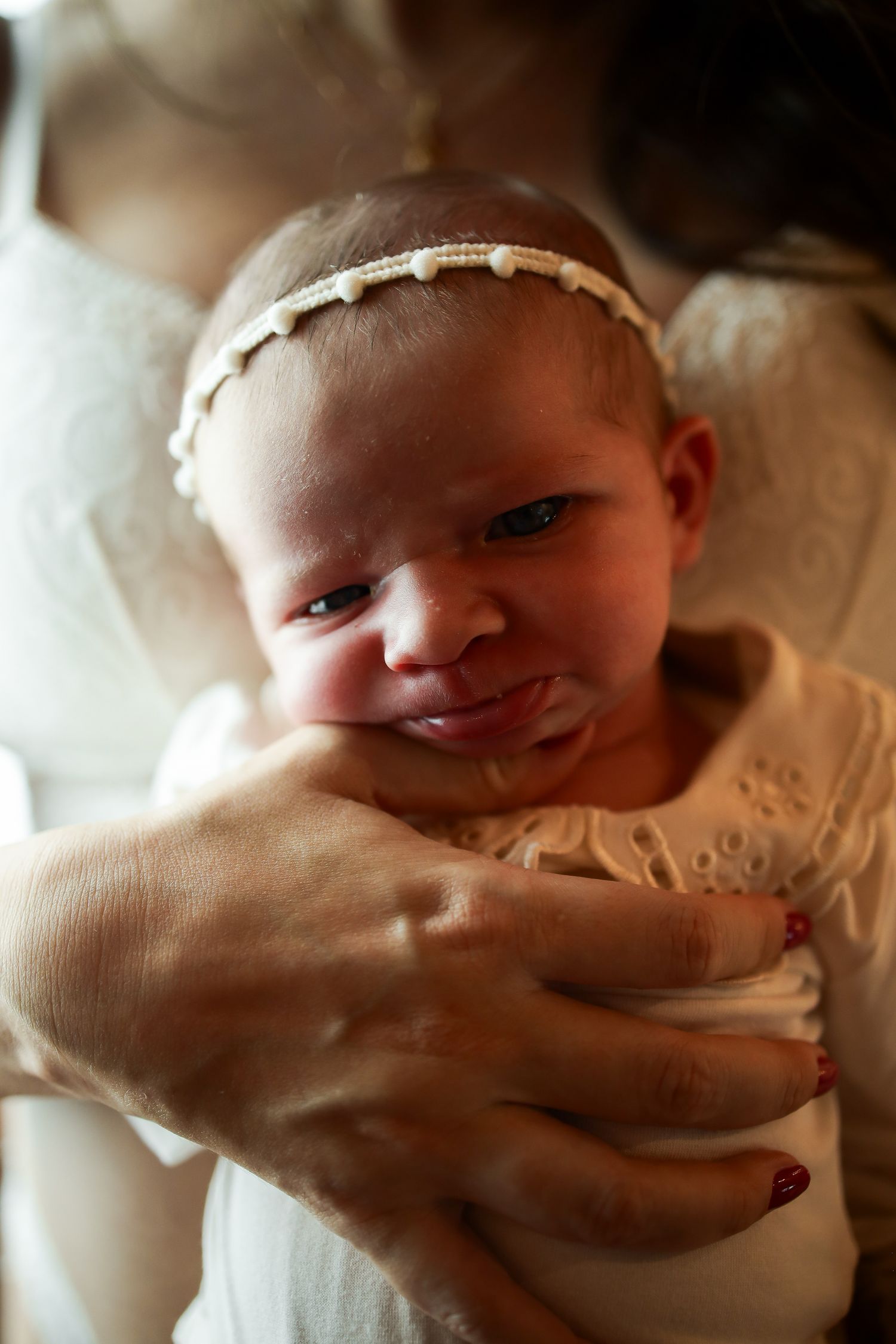 A small baby wearing a delicate white headband smiles while being cradled in someone's hands.