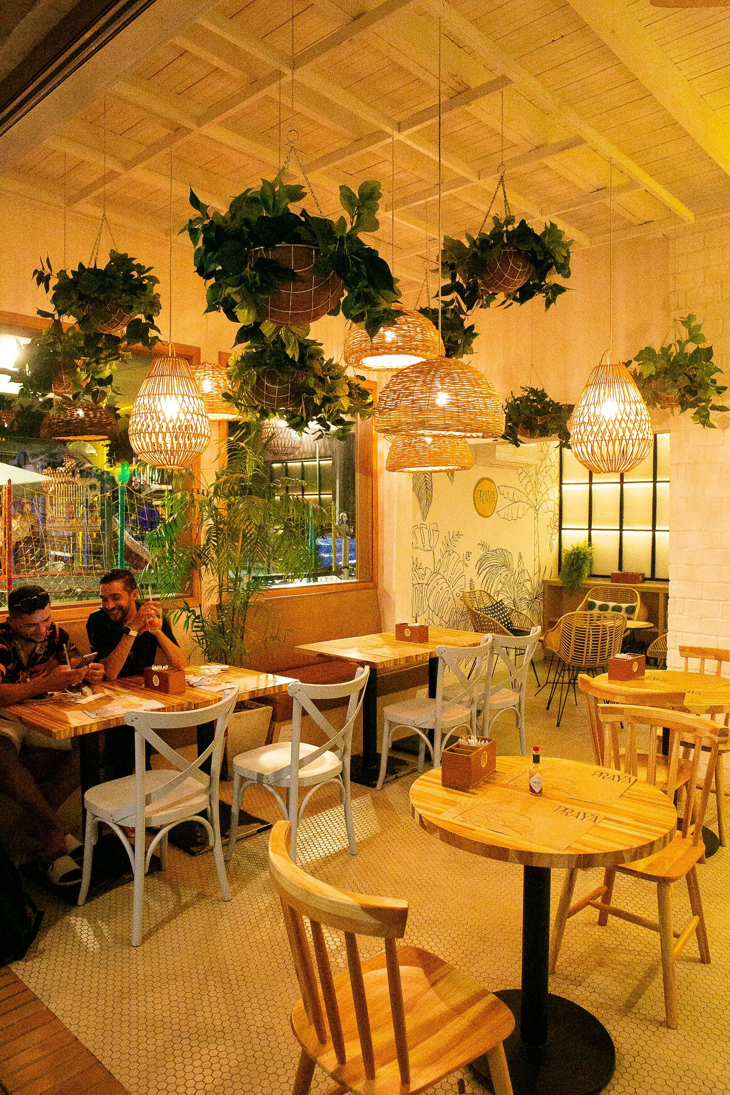 Cozy restaurant interior with hanging wicker lamps, suspended plants, and wooden furniture in warm ambient lighting.