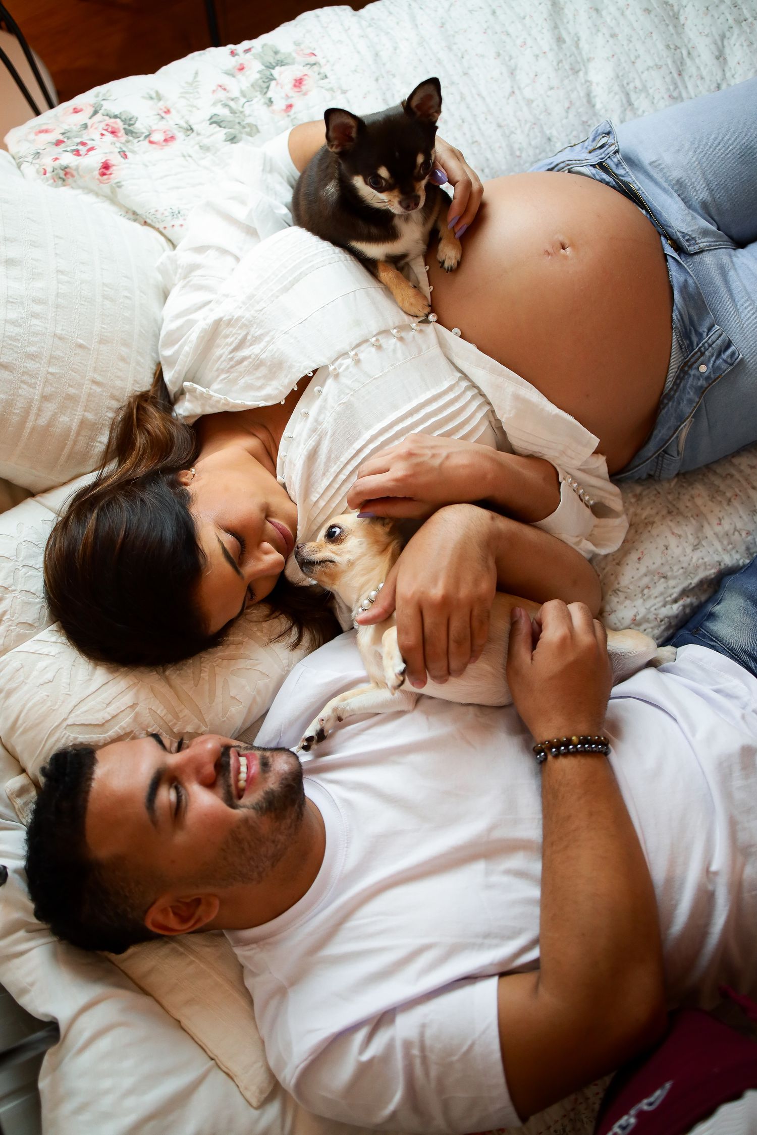 A pregnant couple relaxes in bed with their two cats as pets curl up beside them for a cozy family moment.