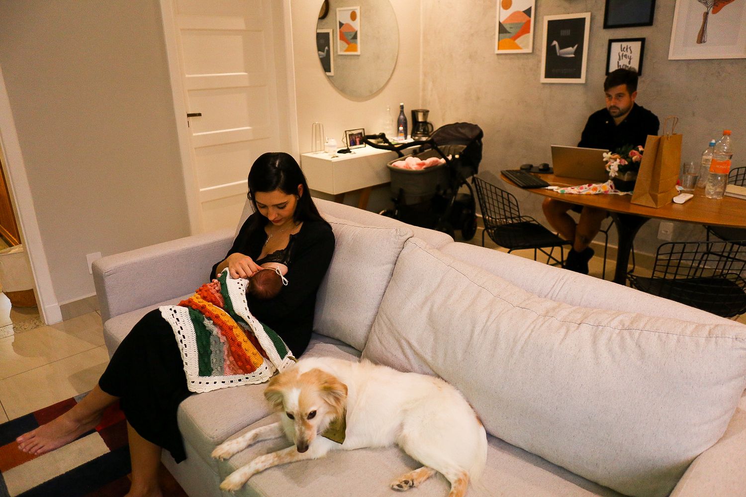 Person relaxing on a light gray couch with a white dog while another person works at a desk in the background.