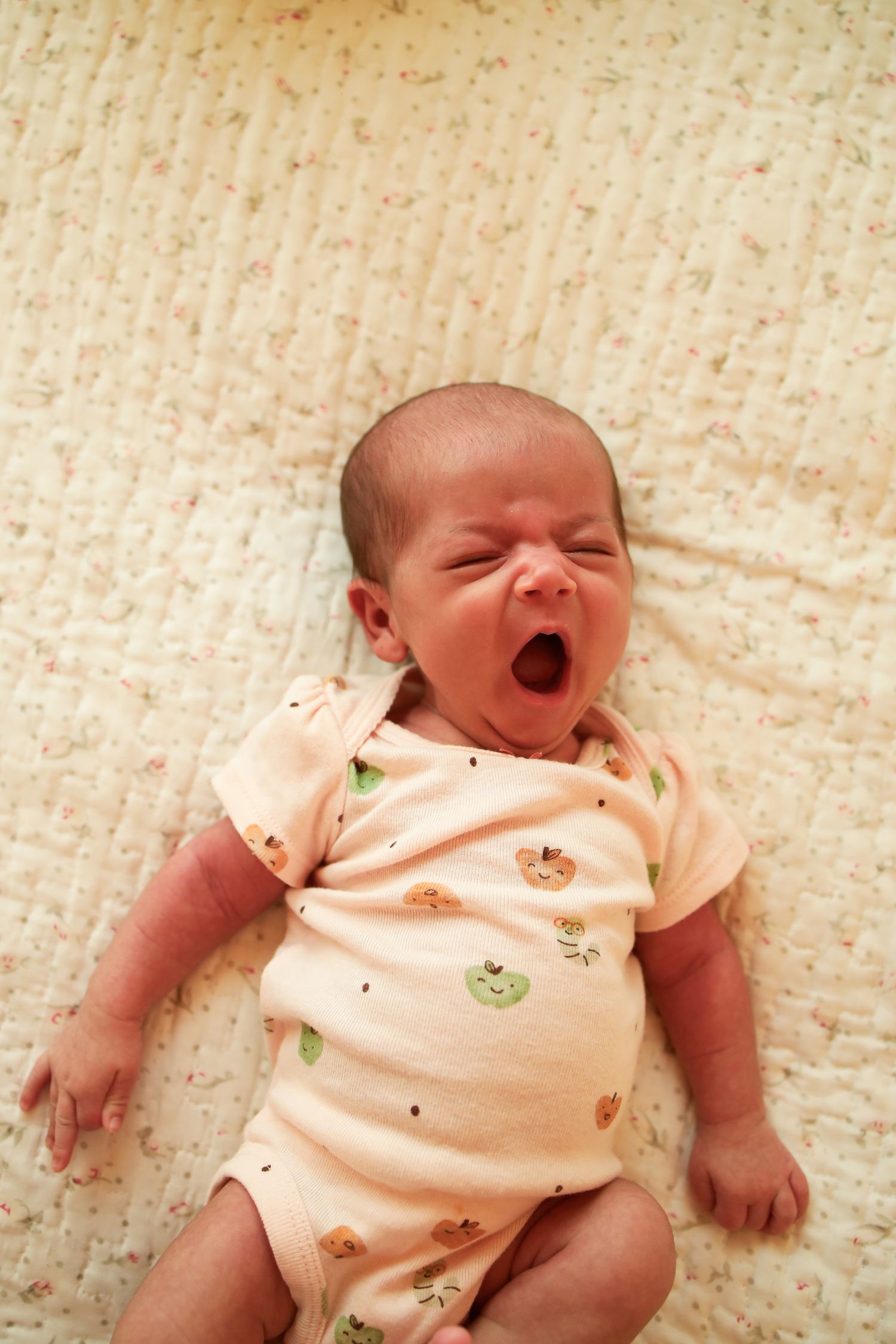 A tiny baby in a peach-colored onesie with fruit prints yawns while lying on a cream-colored textured blanket.
