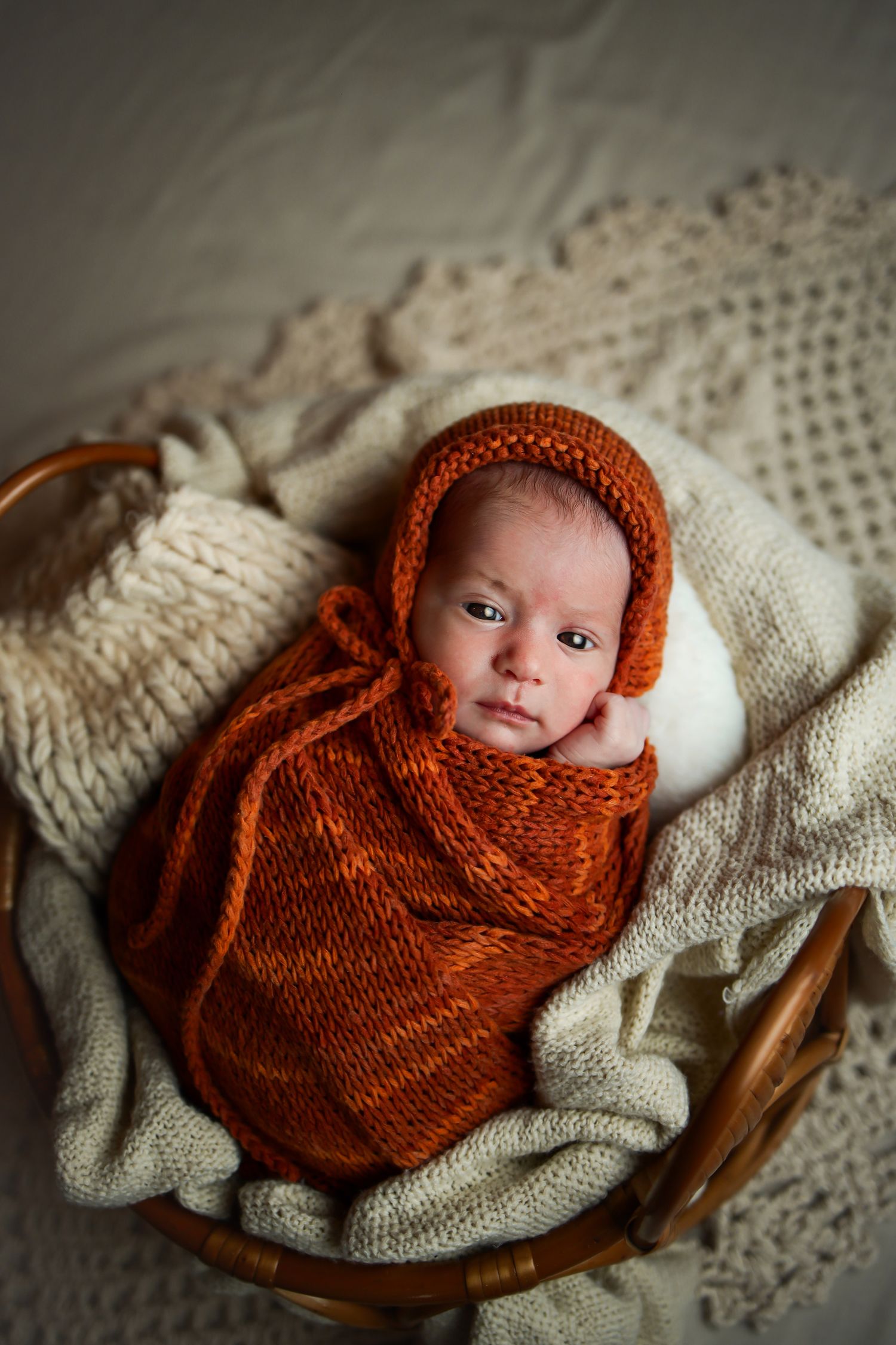 A baby wrapped in a rust colored knit blanket and bonnet lies in a wooden basket on cream textiles.