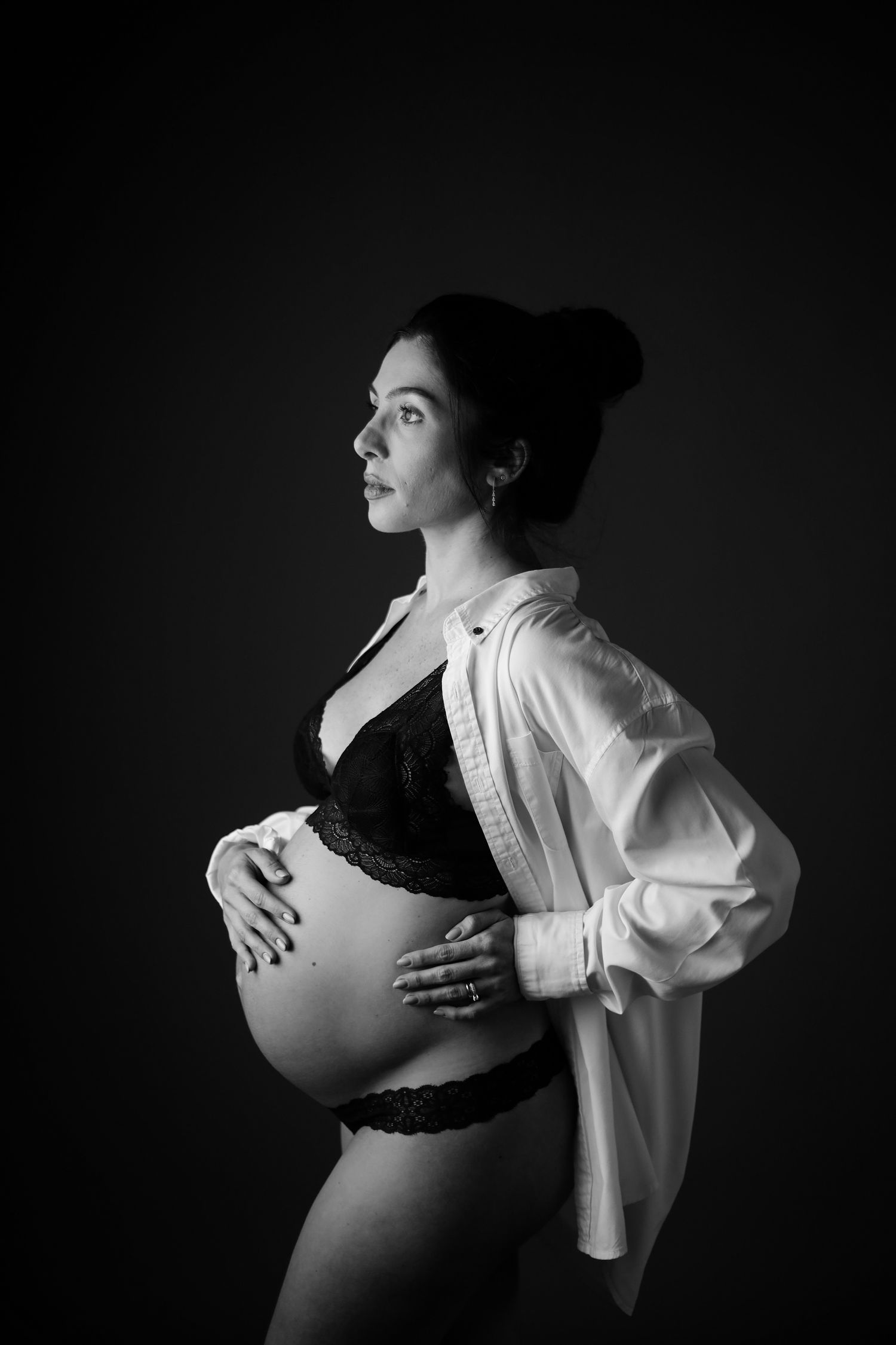 Artistic black and white maternity photo showing figure in lace lingerie and white button-down shirt against dark background.