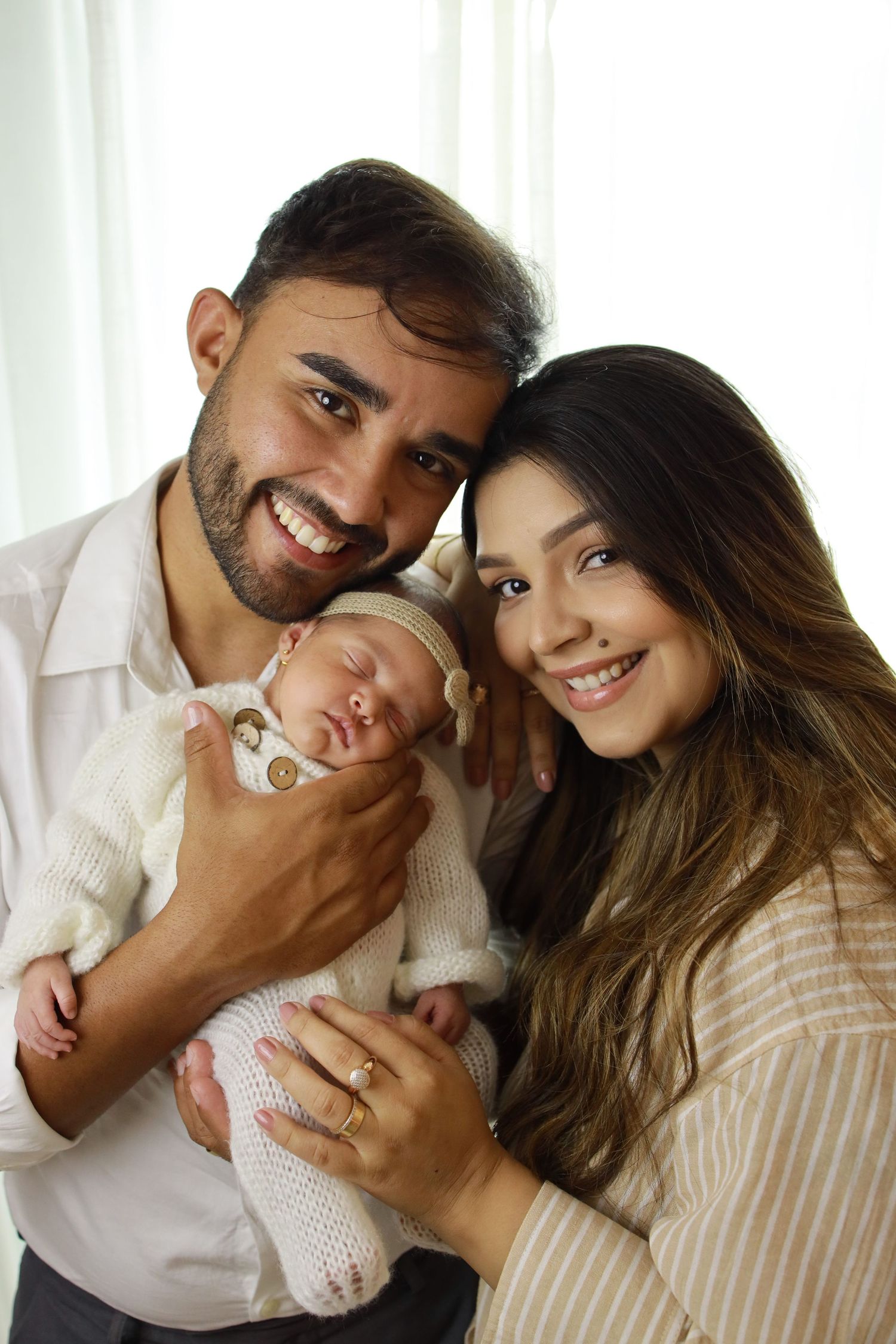 A couple holds their sleeping newborn baby while smiling warmly at the camera.