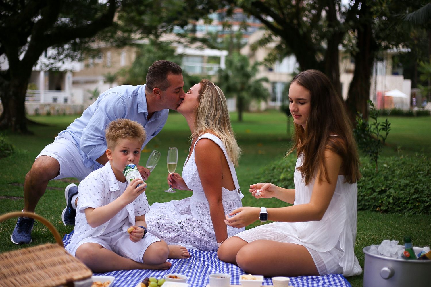 A family enjoys a summer picnic together on a blanket in a park setting, sharing food and laughter outdoors.