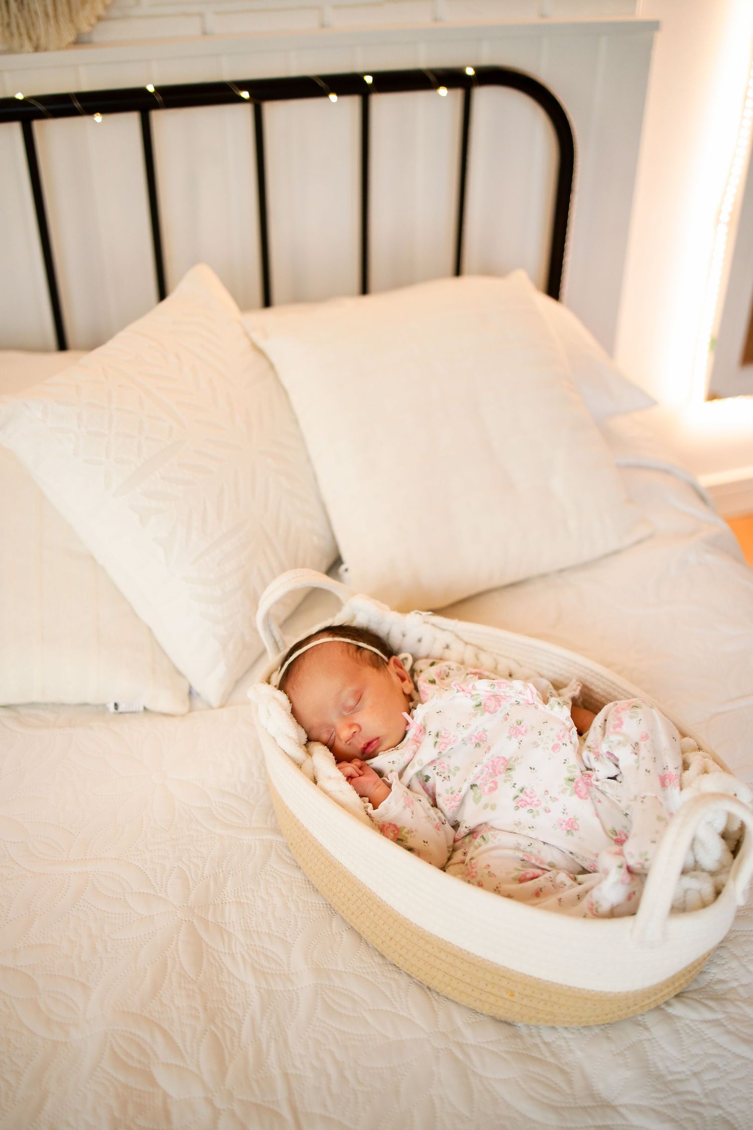 A peaceful newborn sleeping in a cozy rope basket on a bed with white pillows and a black metal headboard.