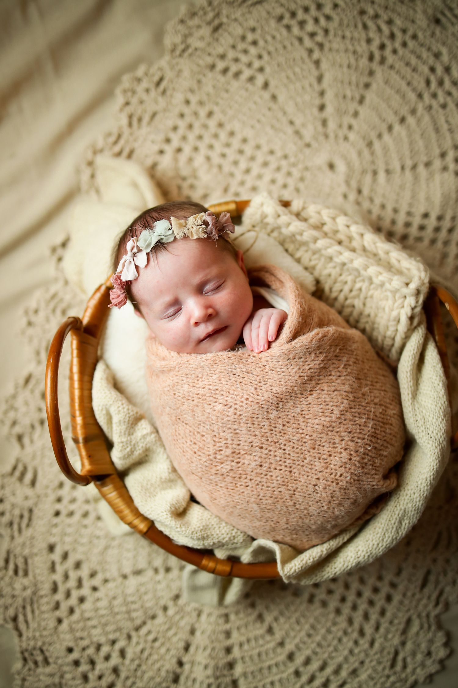 Sleeping newborn swaddled in peach wrap rests peacefully in wooden basket on cream lace blanket.