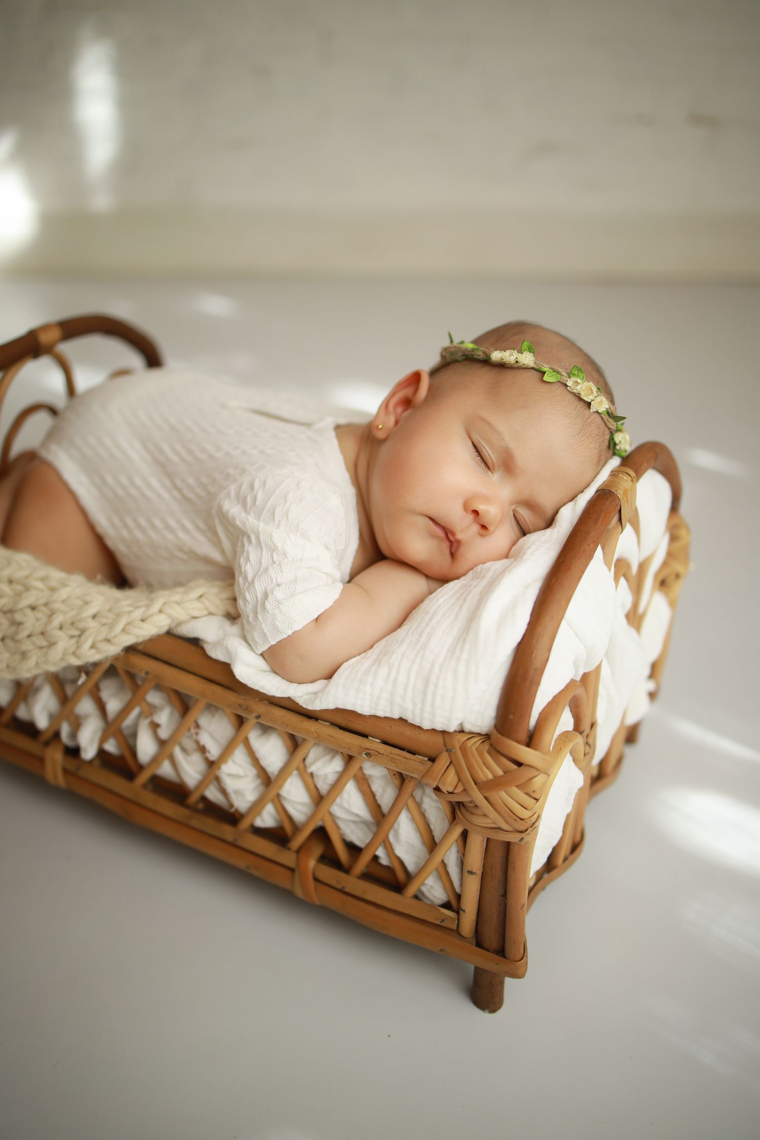 Sleeping newborn in white outfit rests peacefully in vintage wicker doll bed with delicate gold headband.