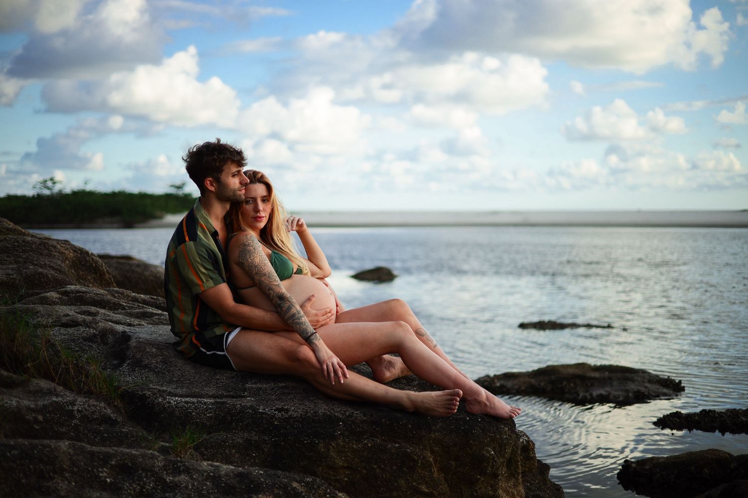 A couple sits together on rocky coastline at sunset overlooking calm ocean waters and cloudy sky.