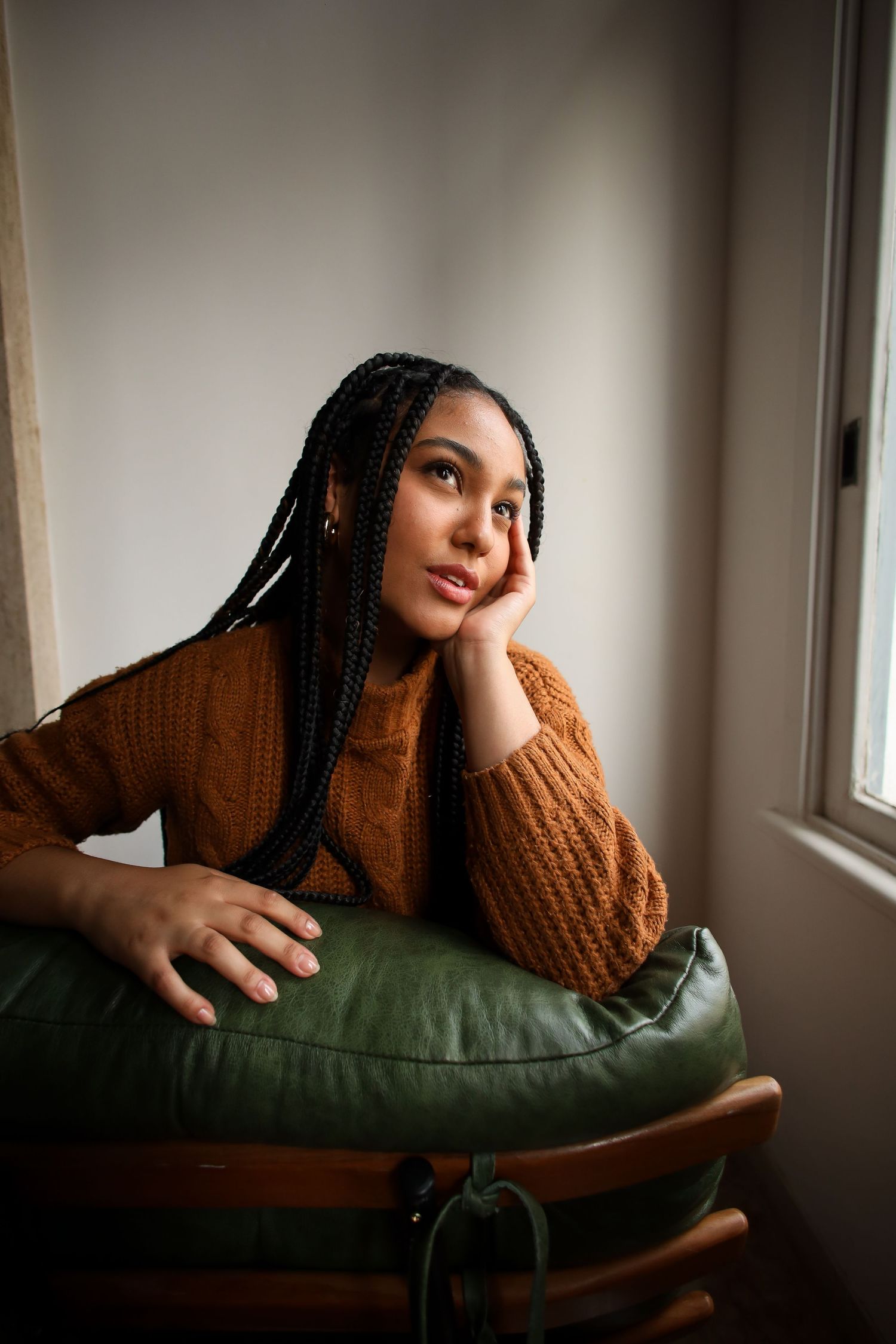 A thoughtful figure in a brown knit sweater rests against a green cushion while gazing out a nearby window.