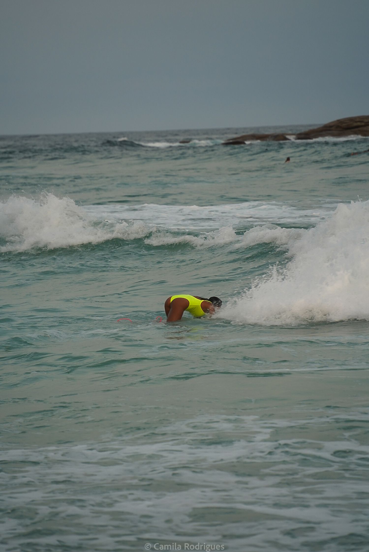 A surfer in bright yellow gear dives under a crashing wave in choppy ocean waters on a cloudy day.