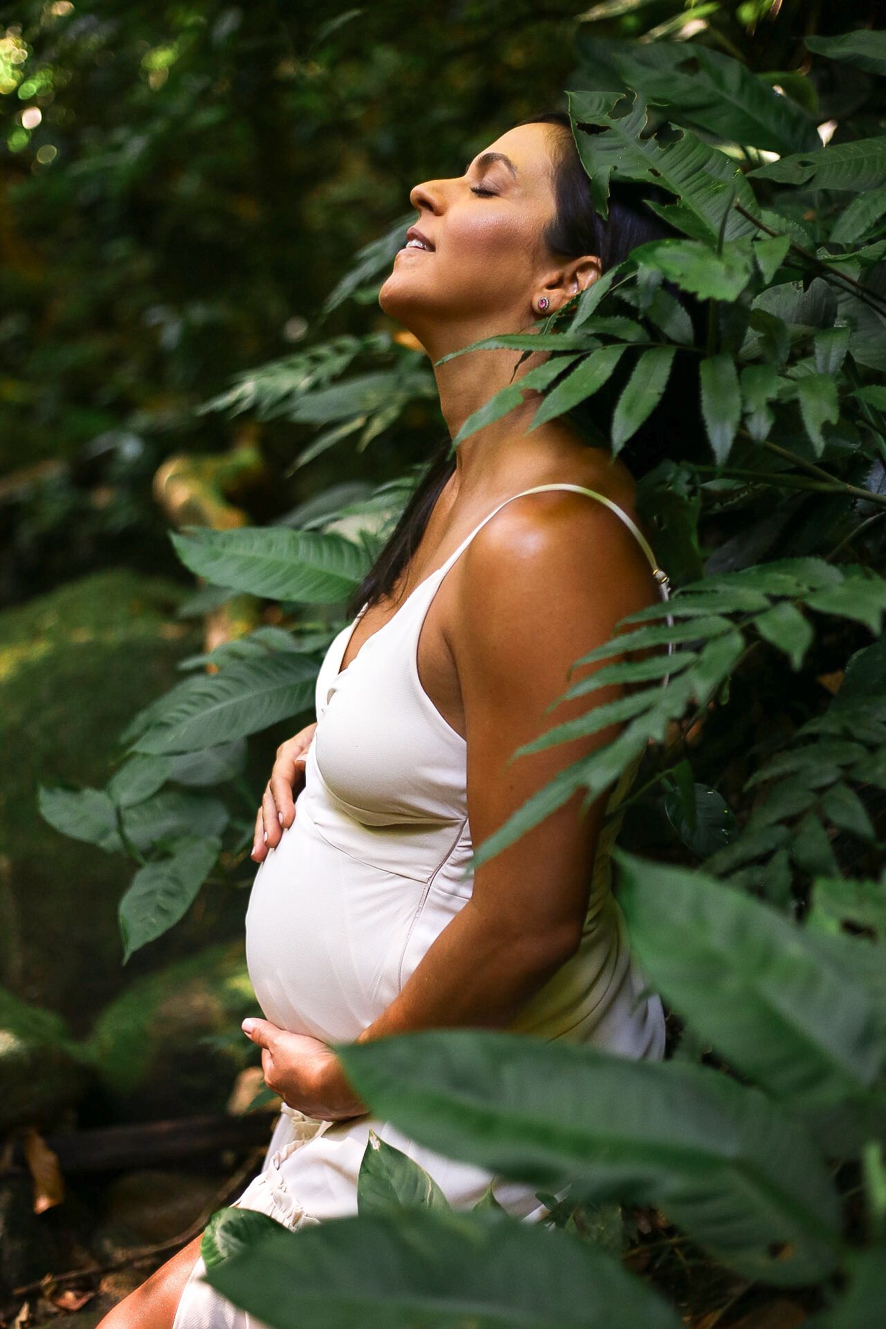Pregnant person in white dress leans back peacefully among green leaves in nature setting.