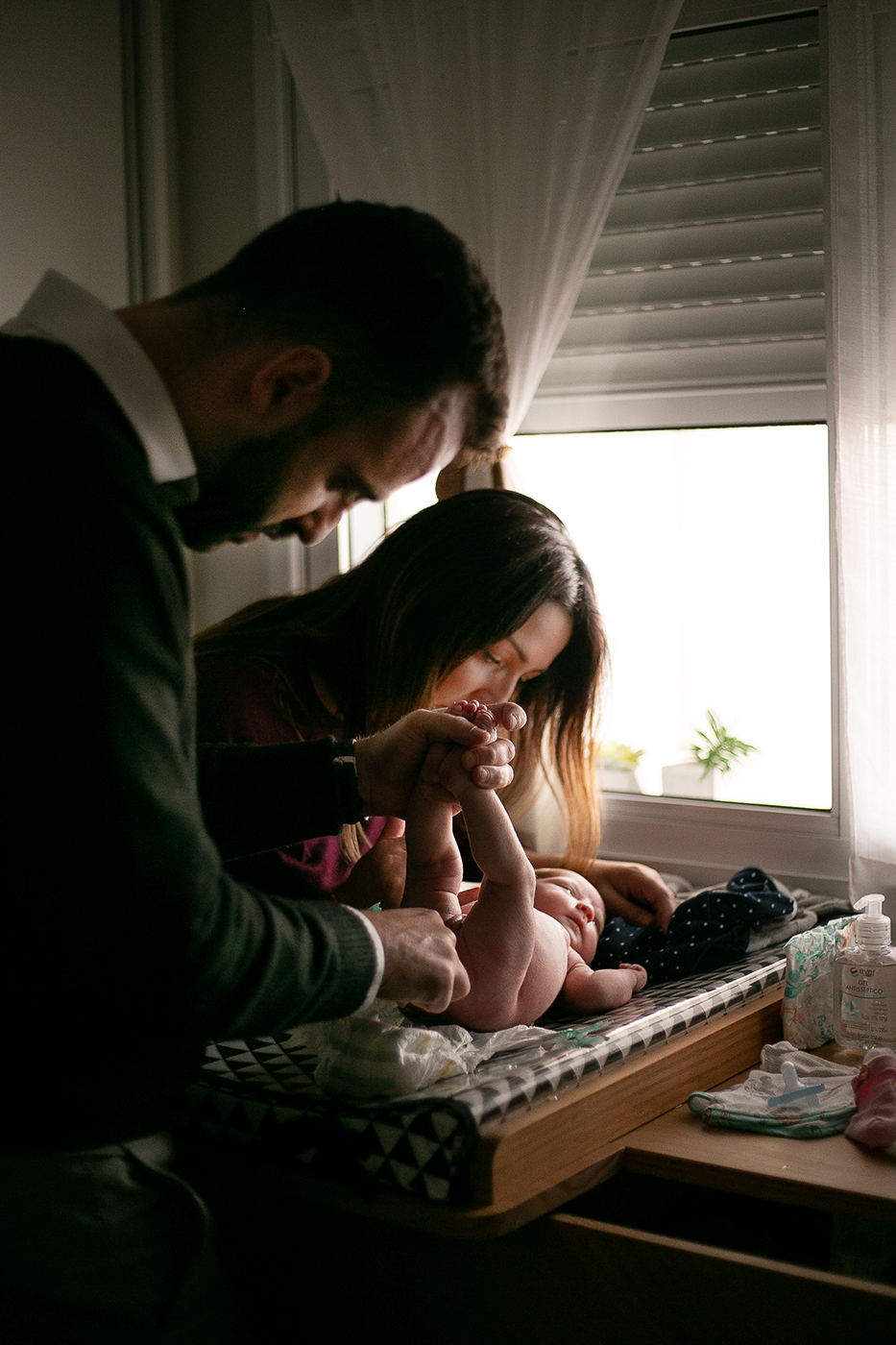 Two people leaning over a changing table in a soft-lit room with a window, sharing an intimate family moment.