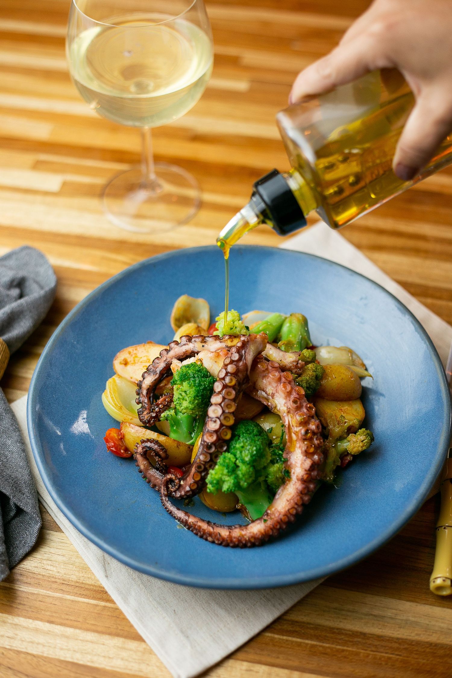 Grilled octopus tentacles served with broccoli and potatoes on blue plate while olive oil is being drizzled on top.