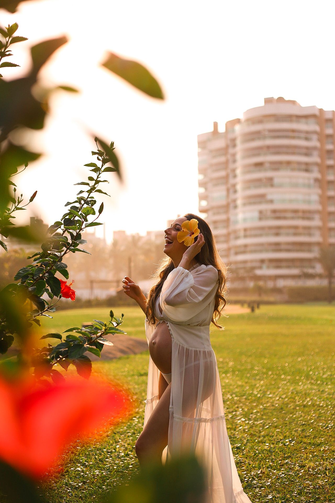 A pregnant woman in a flowing white dress poses at sunset in a garden with red flowers and a high-rise building in background.