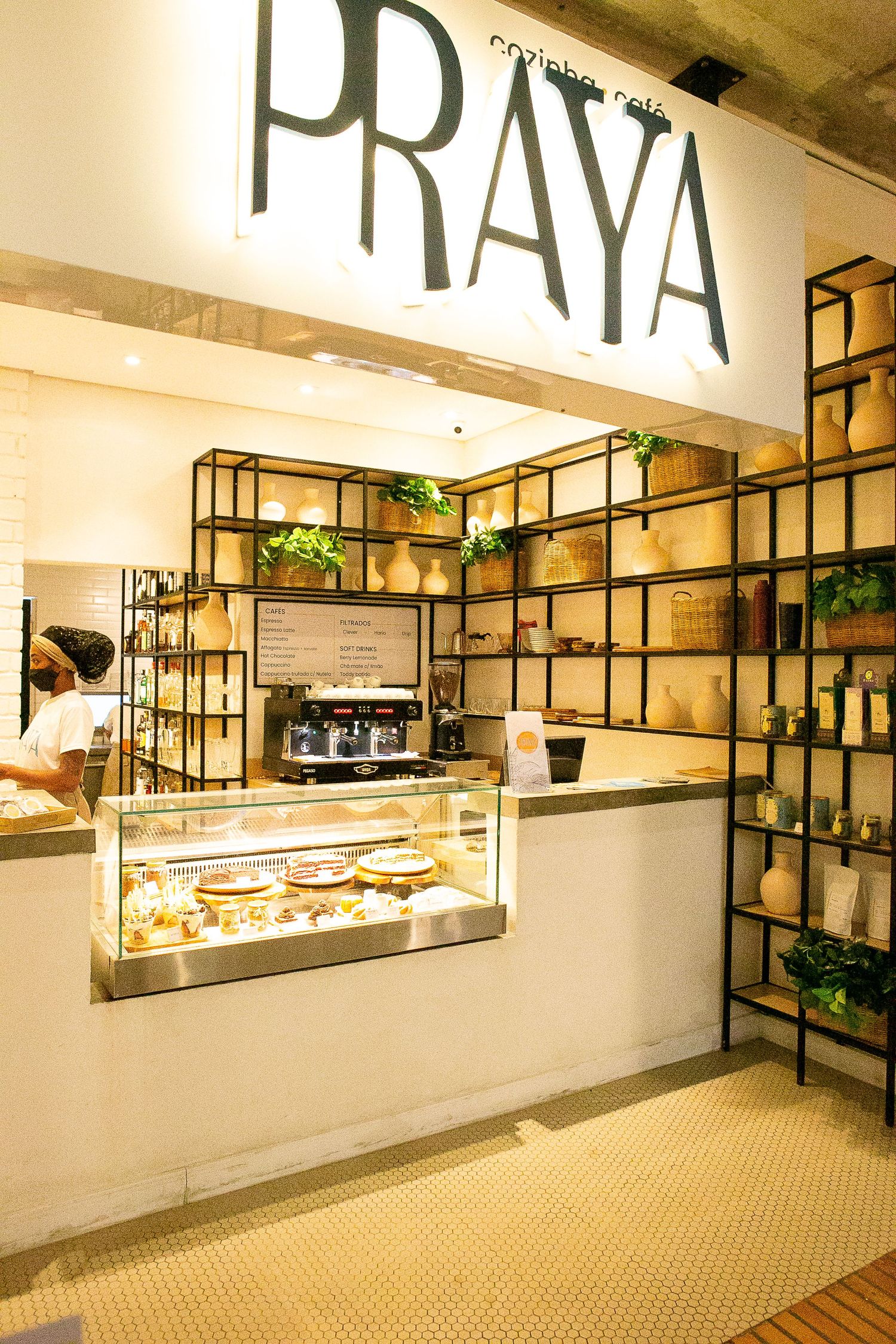 Modern cafe interior with PRAYA signage, industrial shelving, plants, and glass display case in a minimalist white space.