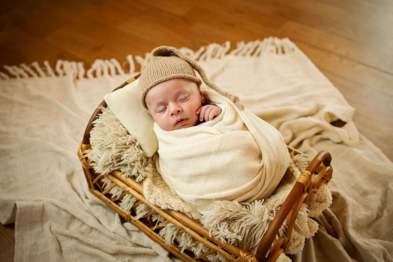 Sleeping newborn baby wrapped in cream blanket wearing beanie hat rests in vintage wicker basket on beige fabric.