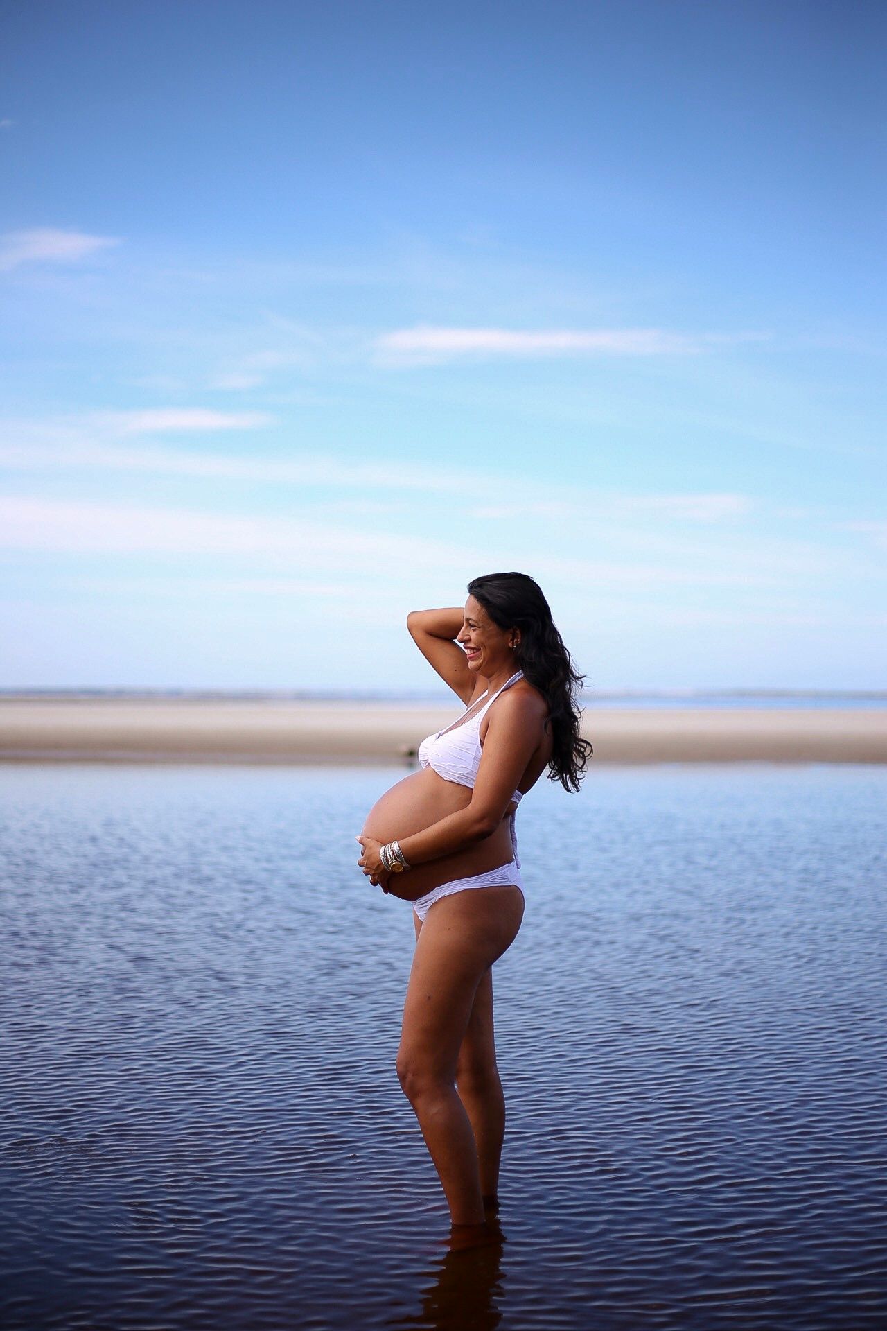 A silhouetted person stands in shallow water against a bright blue sky during a maternity photoshoot at the beach.