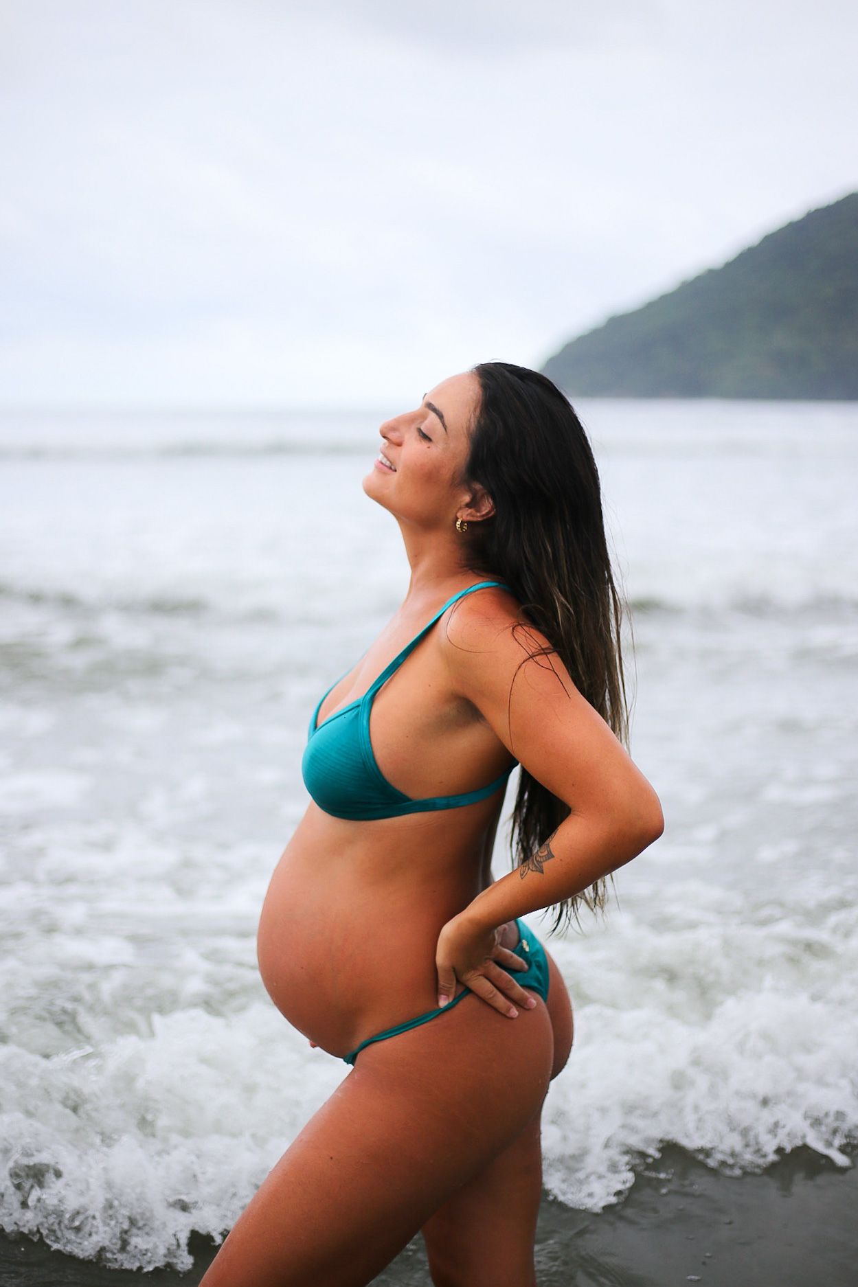 Expectant mother in turquoise bikini stands at ocean's edge with eyes closed and head tilted back serenely.
