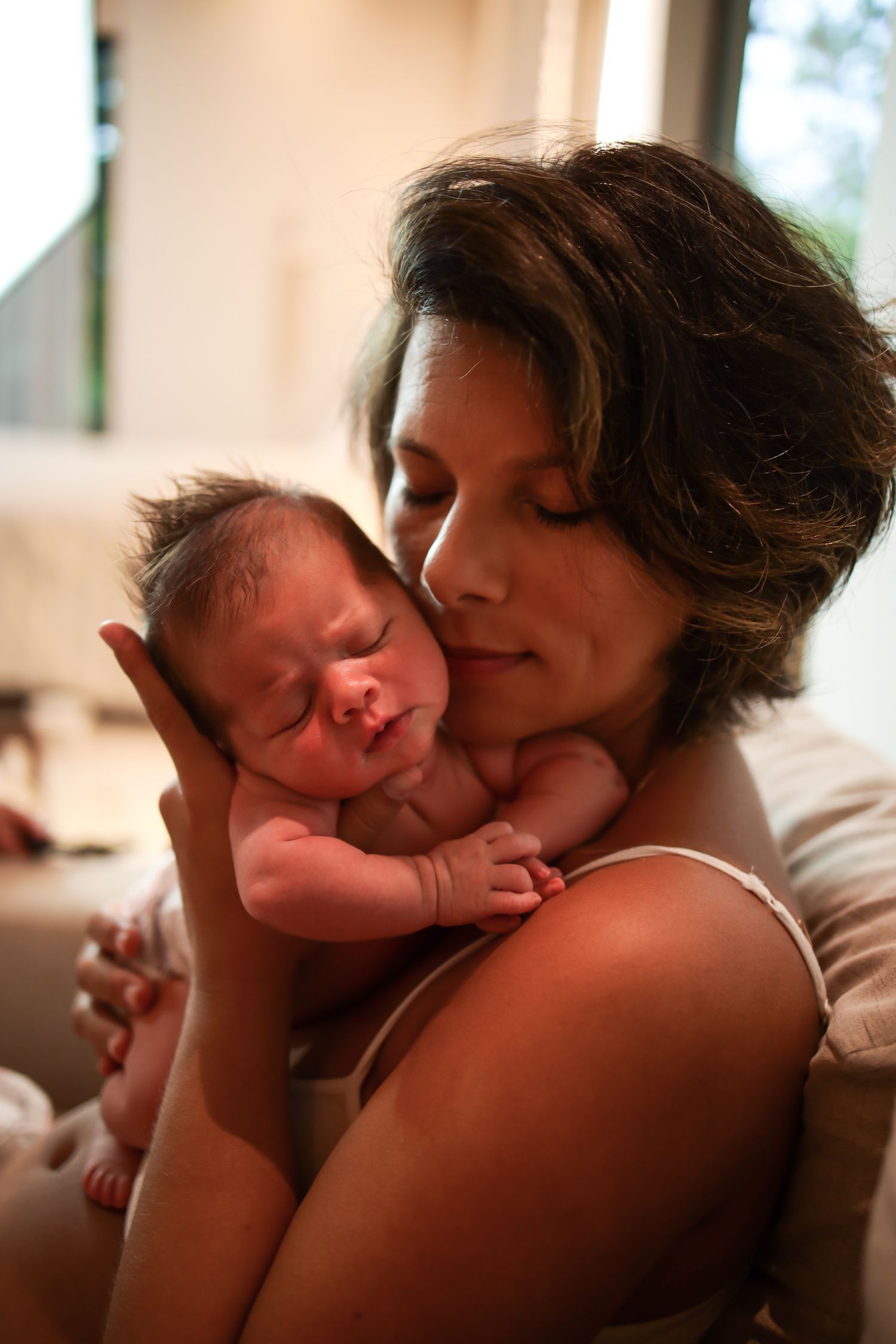 A tender moment between mother and newborn baby in warm natural lighting as they share a close embrace.