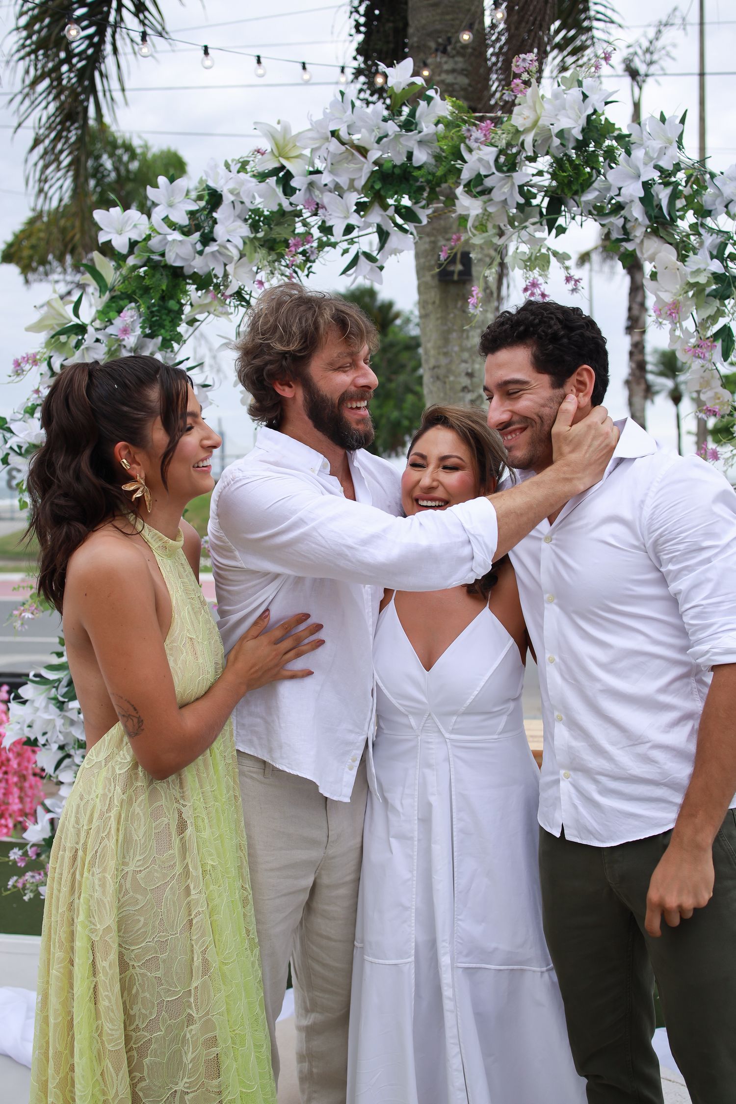 Four people share a joyful moment under a white floral arch decorated with string lights and palm trees.